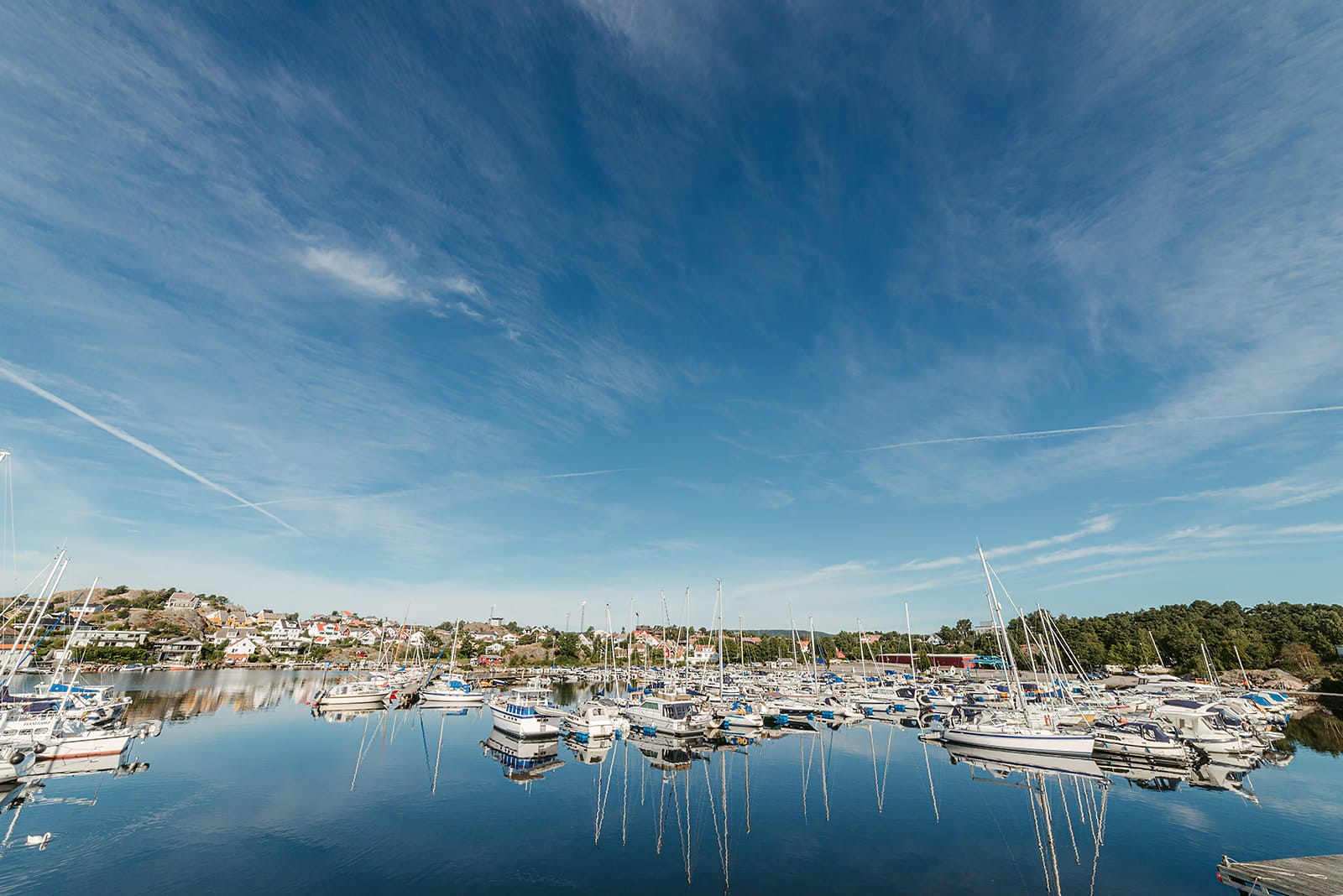 A calm marina near Kristiansand filled with numerous sailboats and yachts reflects a clear blue sky with wispy clouds, bordered by a small coastal town and green trees, in Norway.
