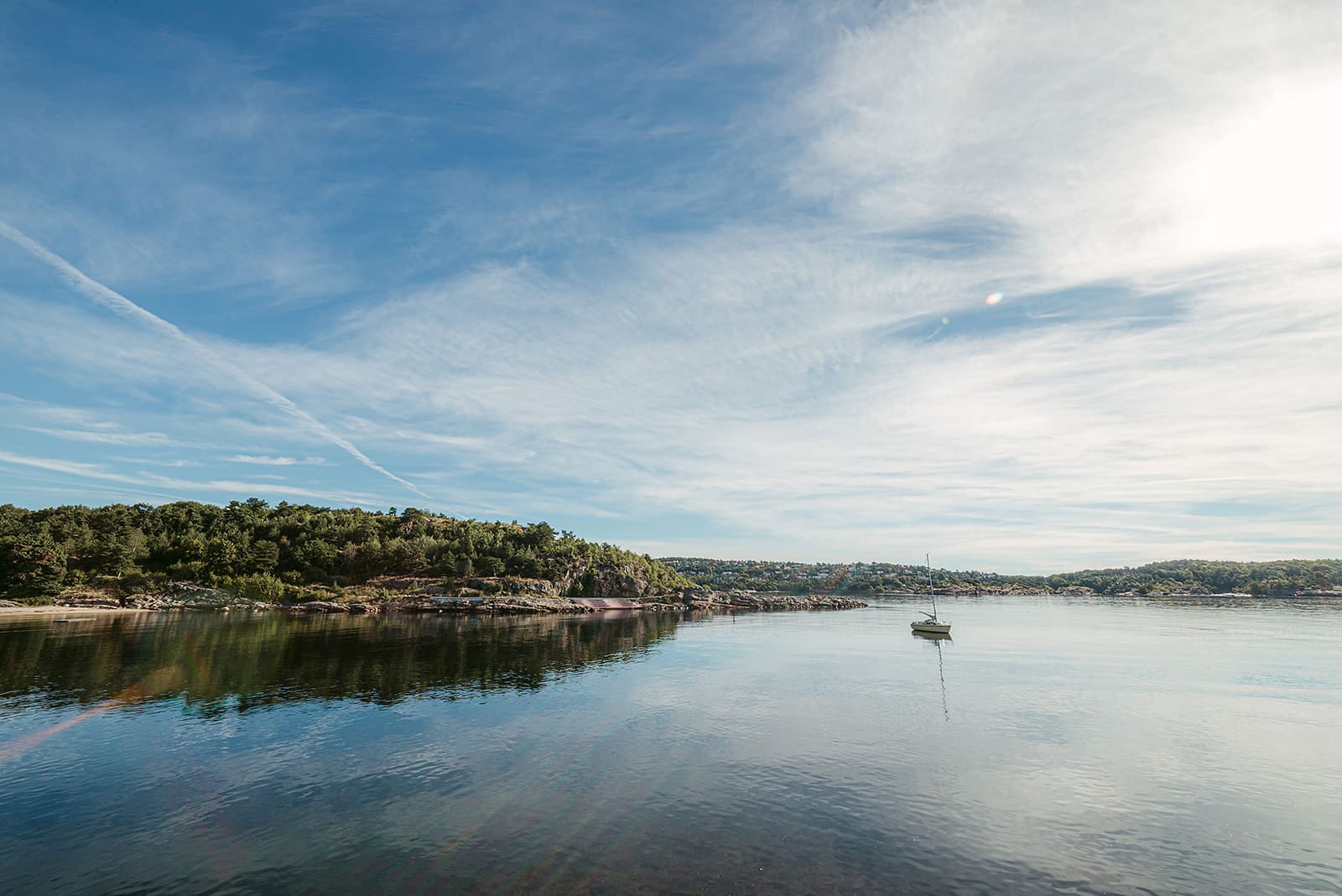 A single sailboat floats on calm water reflecting a partly cloudy sky, surrounded by green, tree-covered hills and a distant shoreline, in Norway.