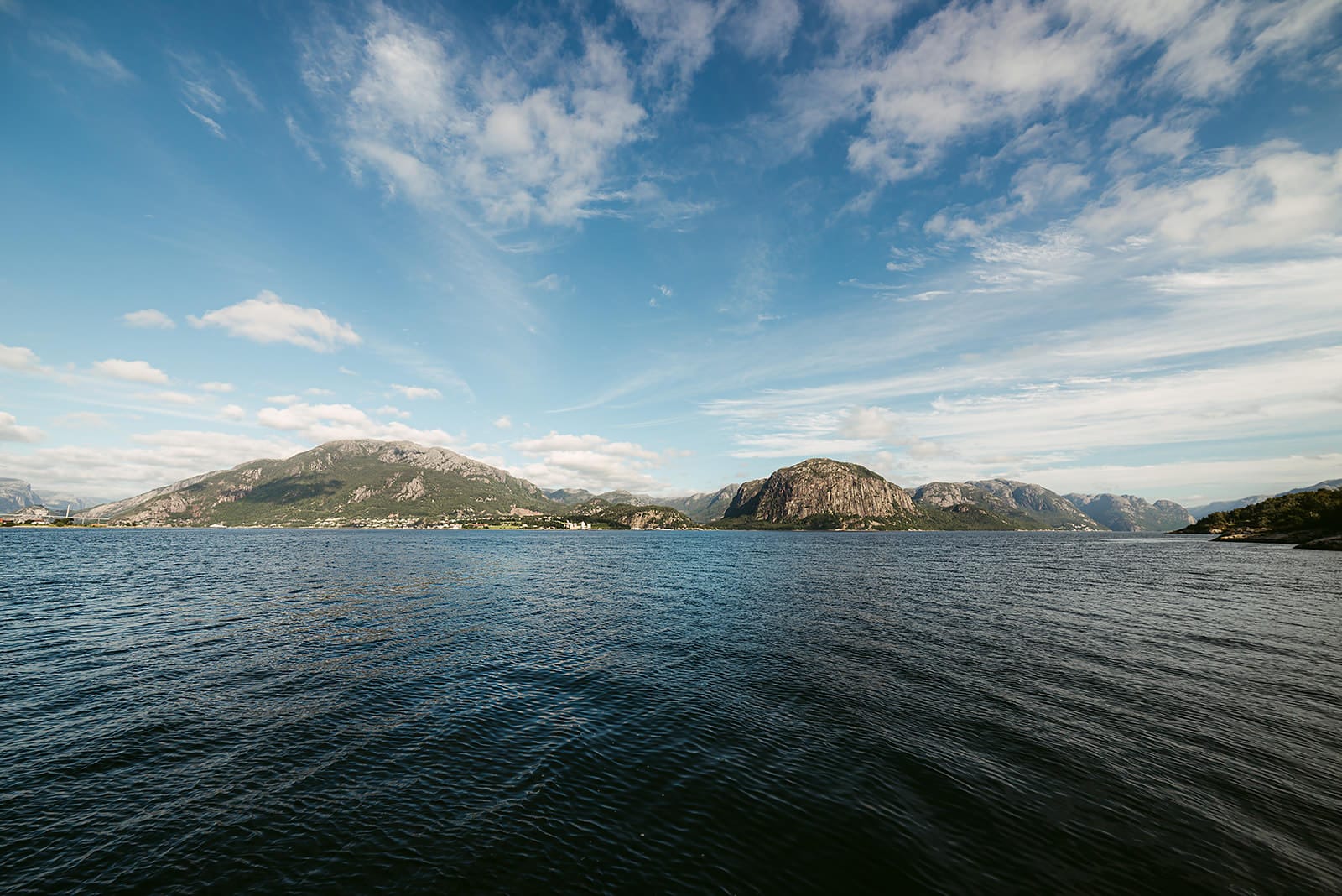 A wide expanse of deep blue water stretches toward rugged, green-covered mountains under a partly cloudy sky, capturing a scenic fjord landscape in Norway.
