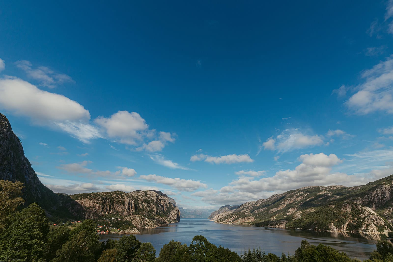 A narrow fjord flanked by steep, rocky mountains with patches of green vegetation extends under a bright blue sky with scattered clouds, showcasing a dramatic natural landscape in Norway.