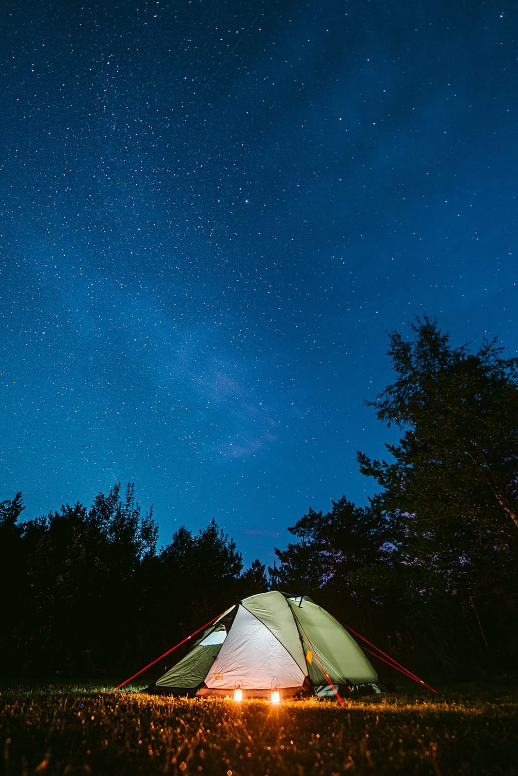 A glowing tent illuminated by two lanterns sits on grass surrounded by trees under a clear, star-filled night sky in Norway.