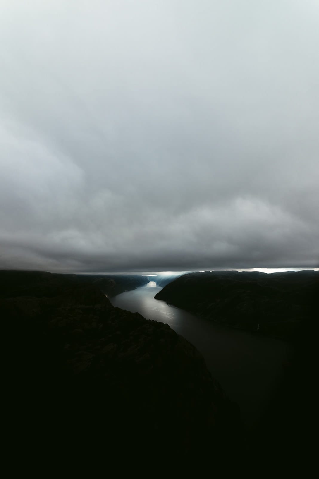 A dramatic fjord landscape with steep, dark mountains on either side is partially illuminated by a narrow beam of light breaking through heavy, low-hanging clouds in Norway.