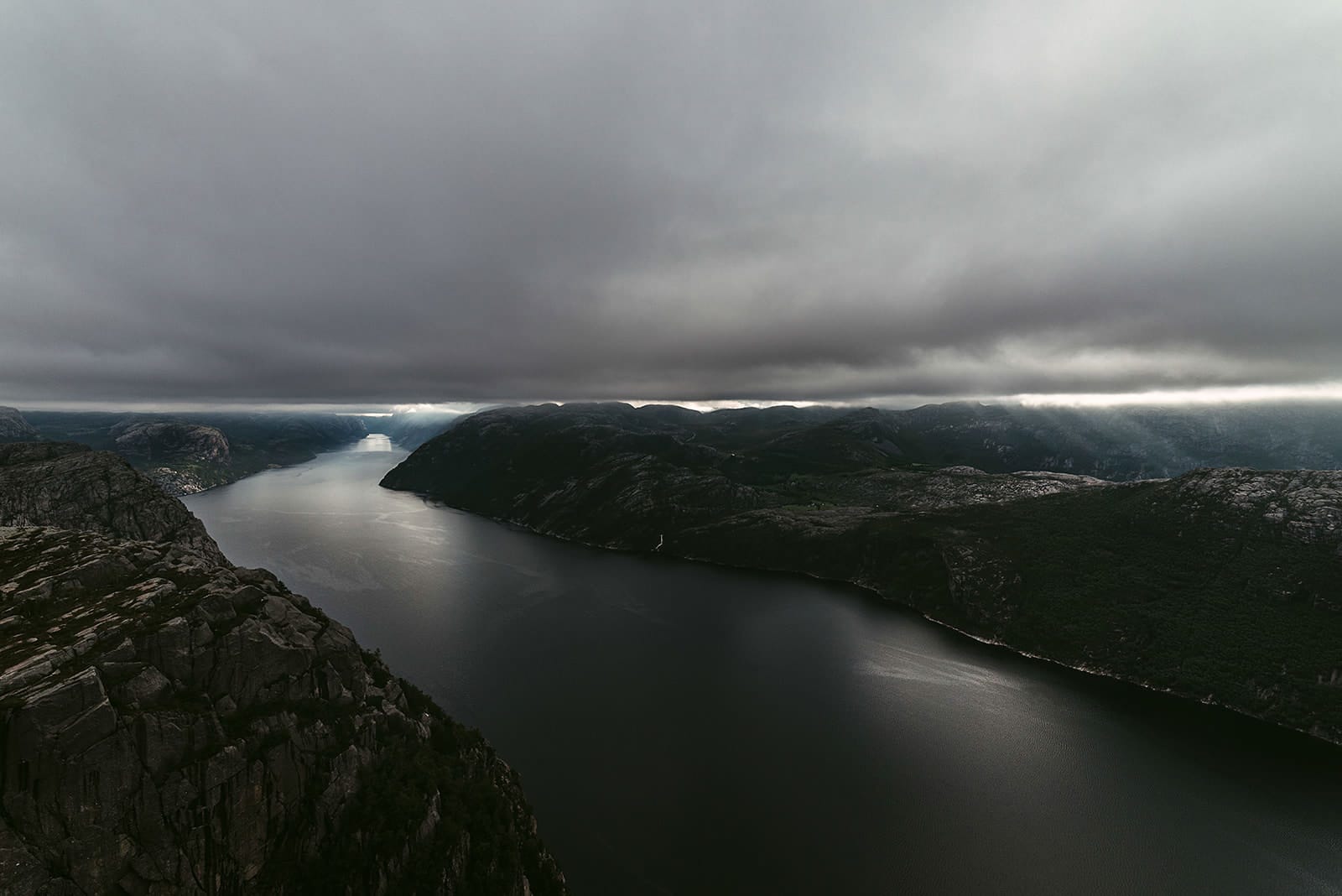 A dark fjord winds between steep, rocky mountains under a thick layer of low clouds, with rays of sunlight breaking through the clouds and illuminating parts of the landscape in Norway.