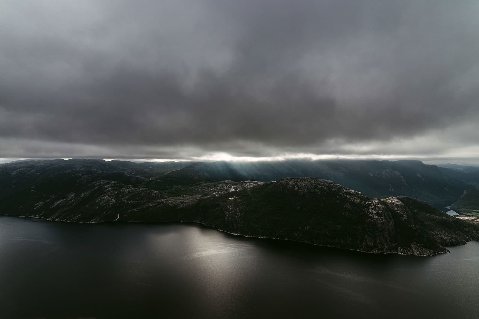A fjord with dark, rocky hills is partially illuminated by rays of sunlight breaking through thick, low-hanging clouds, creating a dramatic contrast in Norway.