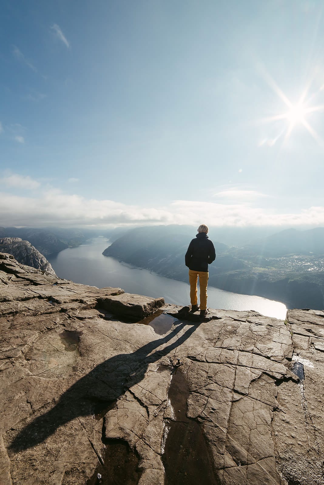 A person stands on a rocky cliff overlooking a sunlit fjord and distant mountains under a clear sky in Norway.