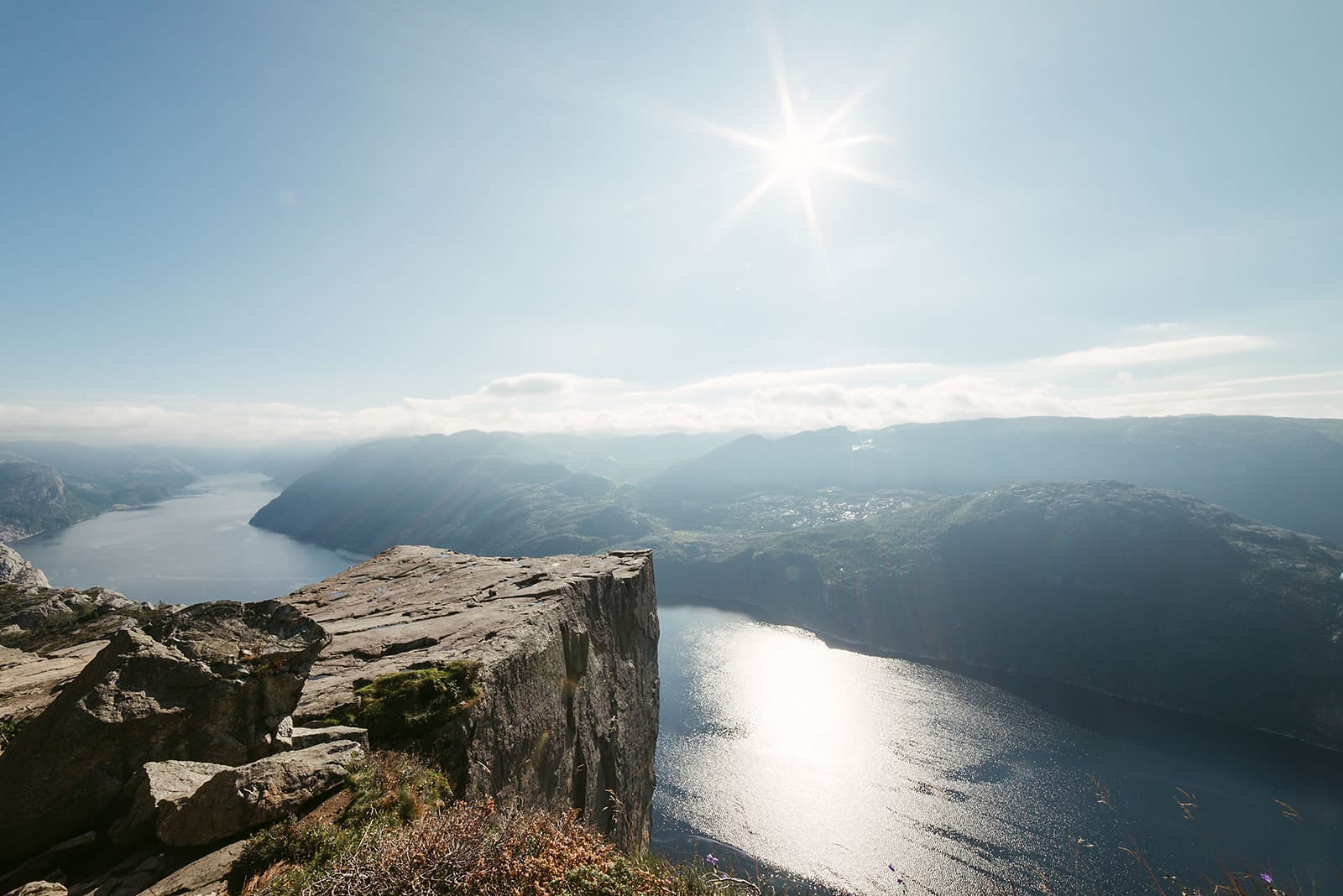 A sunlit cliff juts out over a shimmering fjord surrounded by distant mountains under a clear blue sky, capturing a breathtaking view in Norway.