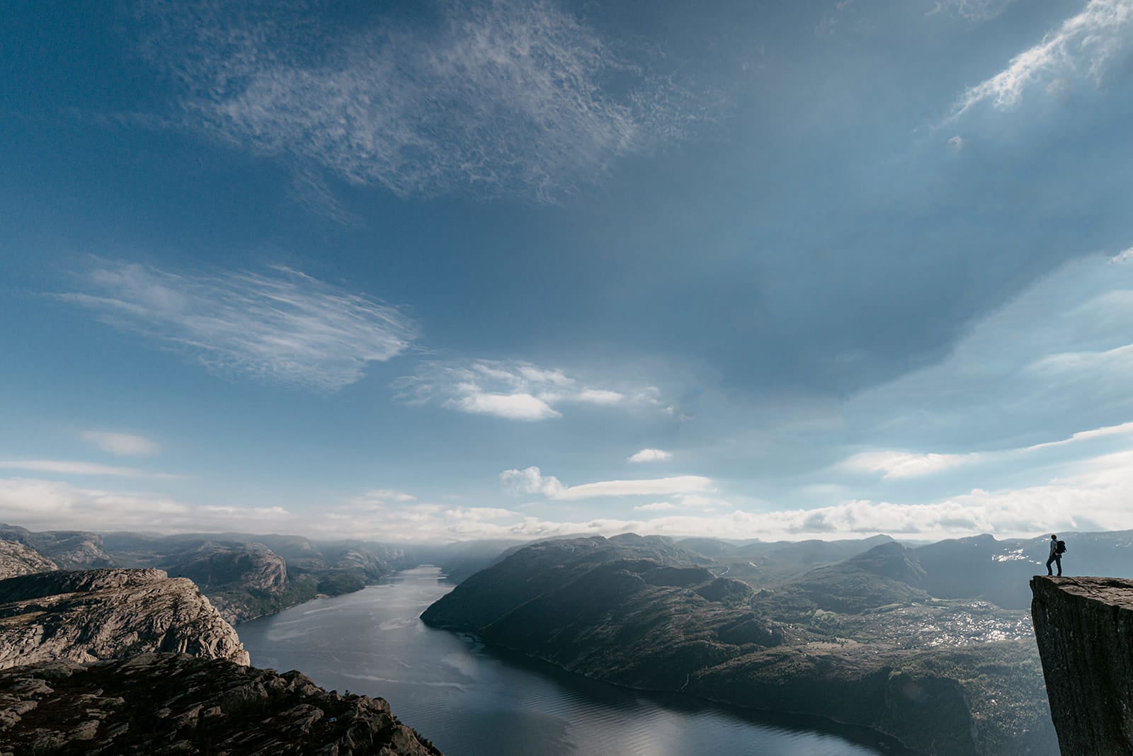 A person stands on the edge of a rocky cliff overlooking a vast fjord and mountainous landscape under a partly cloudy blue sky in Norway.