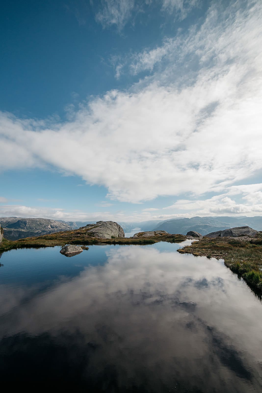 A calm mountain pond reflects the blue sky and clouds, with rocky terrain and distant fjords visible under a partly cloudy sky in Norway.