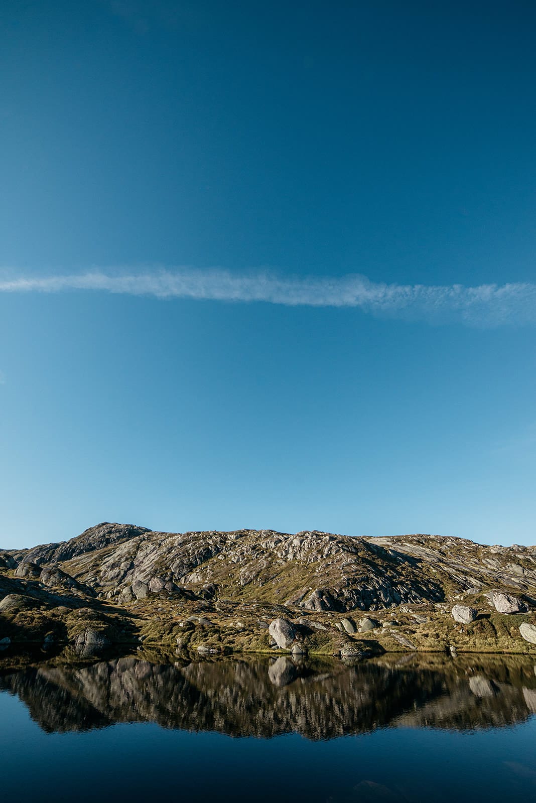 A rocky hillside is reflected in the still water of a mountain pond under a clear blue sky with a single contrail in Norway.