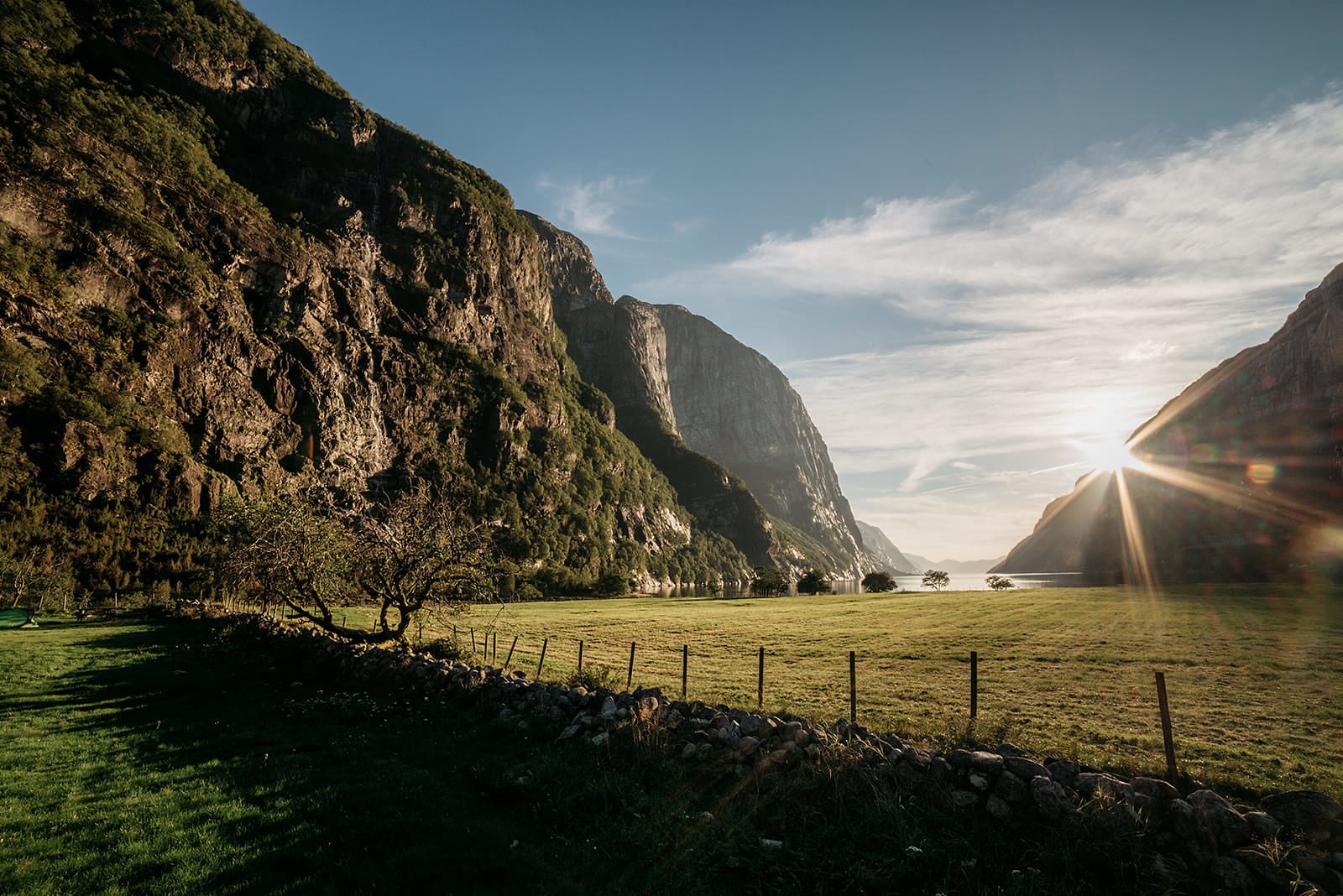 A sunlit valley with a grassy field and scattered trees is framed by steep, rocky cliffs, with the sun setting behind the mountains in Norway.