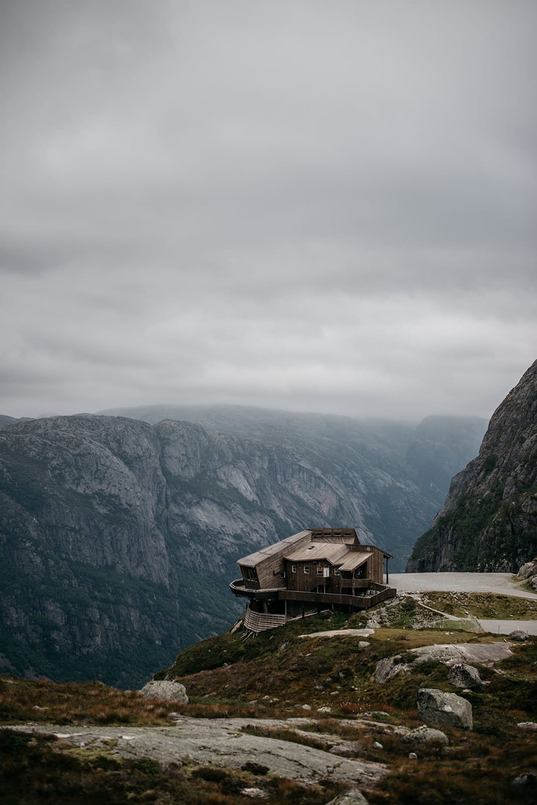 A wooden building perched on a rocky hillside overlooks a deep fjord surrounded by steep, mist-covered mountains under a cloudy sky in Norway.