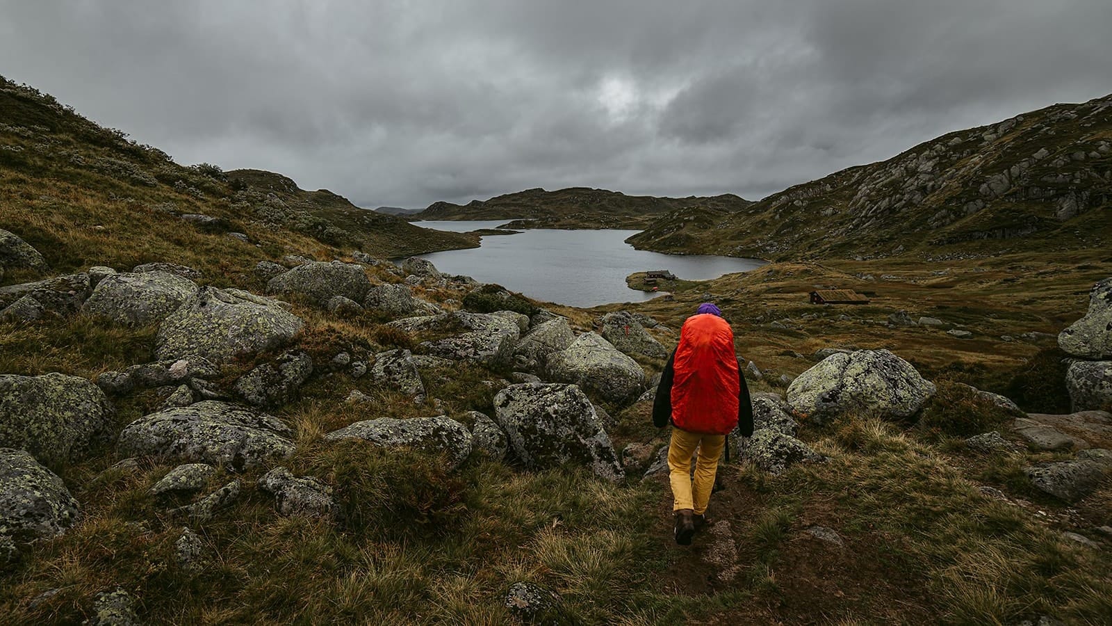 A lone hiker wearing a bright orange backpack and yellow pants walks along a rocky trail toward a serene lake surrounded by rugged hills under a cloudy sky, in a remote area of Norway.