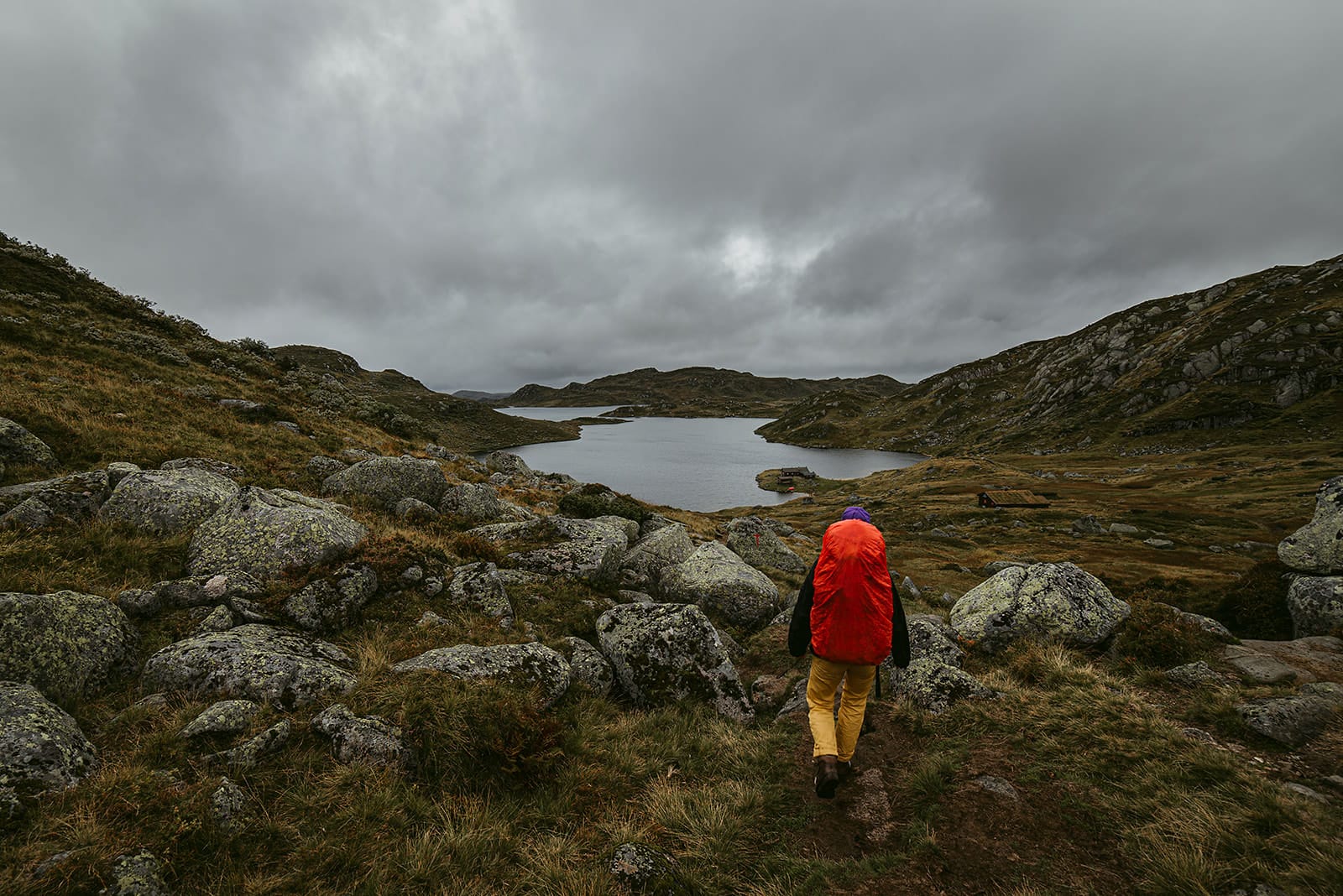 A lone hiker wearing a bright orange backpack and yellow pants walks along a rocky trail toward a serene lake surrounded by rugged hills under a cloudy sky, in a remote area of Norway.