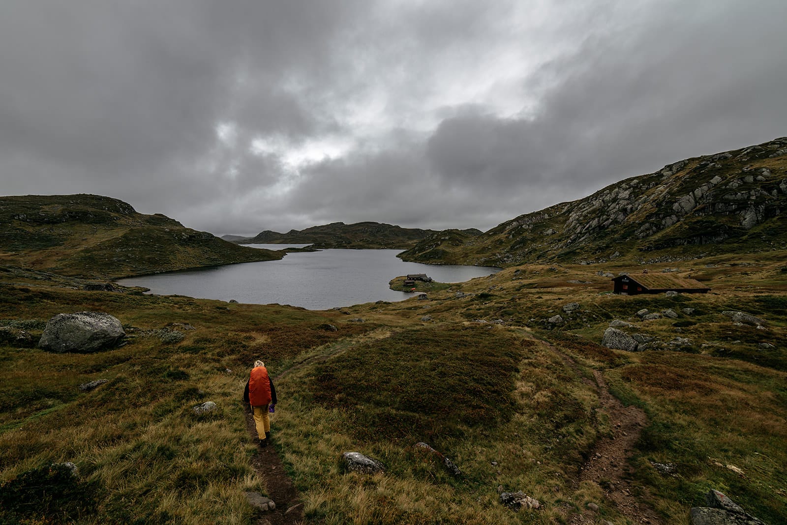 A person with a bright orange backpack walks along a narrow trail through a grassy, rocky landscape toward a lake surrounded by hills under a cloudy sky in Norway.