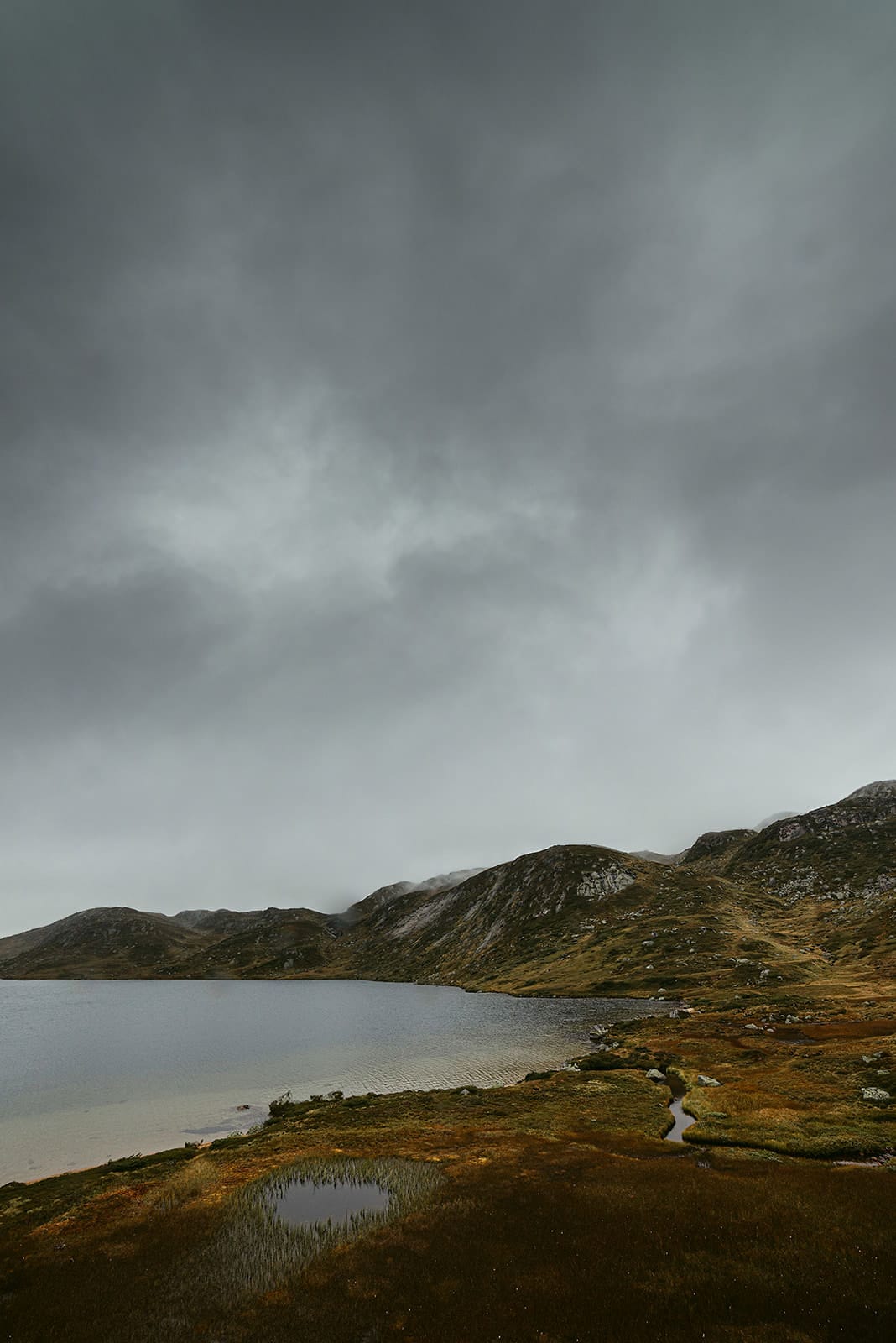 A moody, overcast sky hangs over a calm lake surrounded by rocky hills and grassy terrain in Norway.