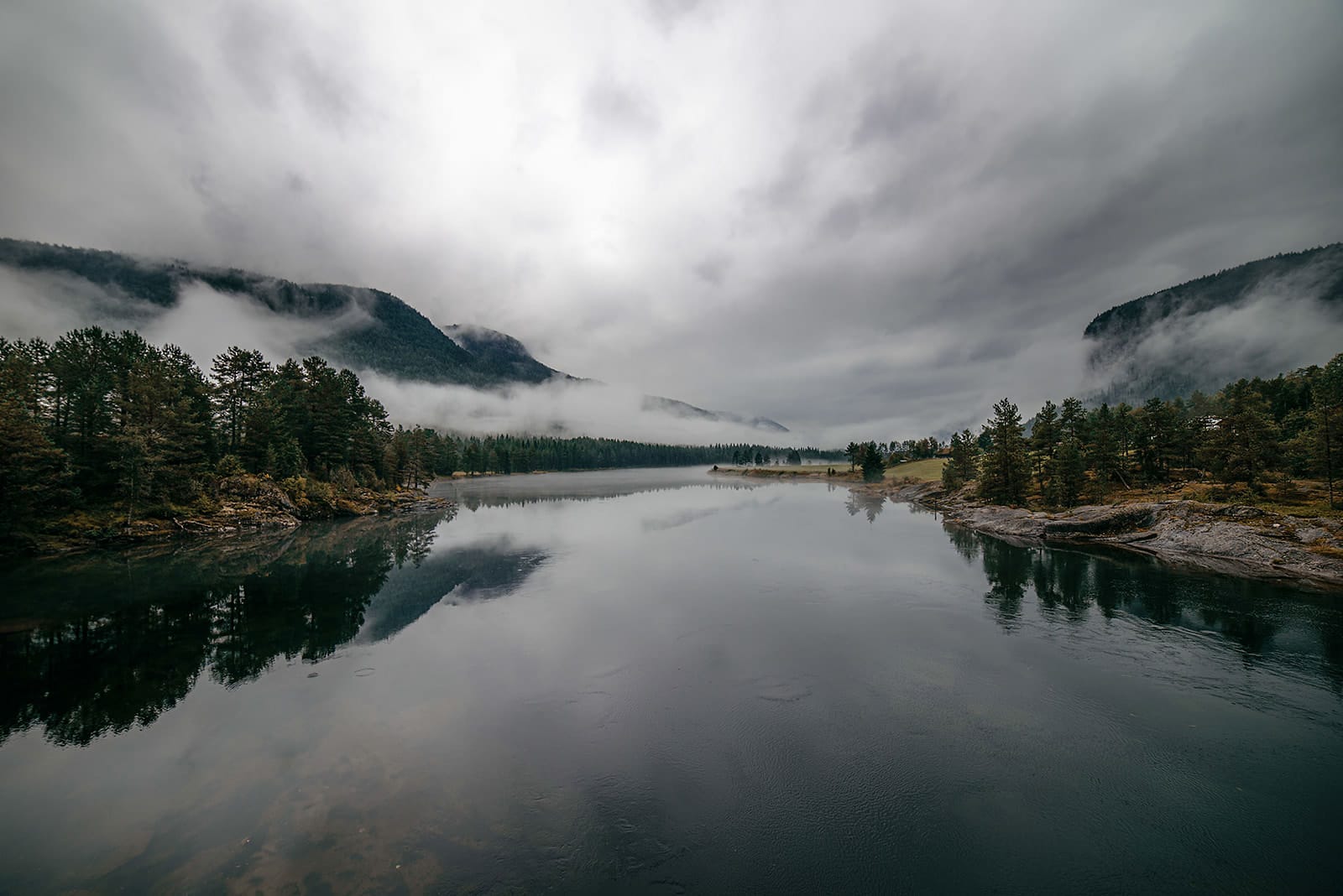 A misty river flanked by dense pine forests reflects the cloudy sky and fog-covered mountains in a serene Norwegian landscape.