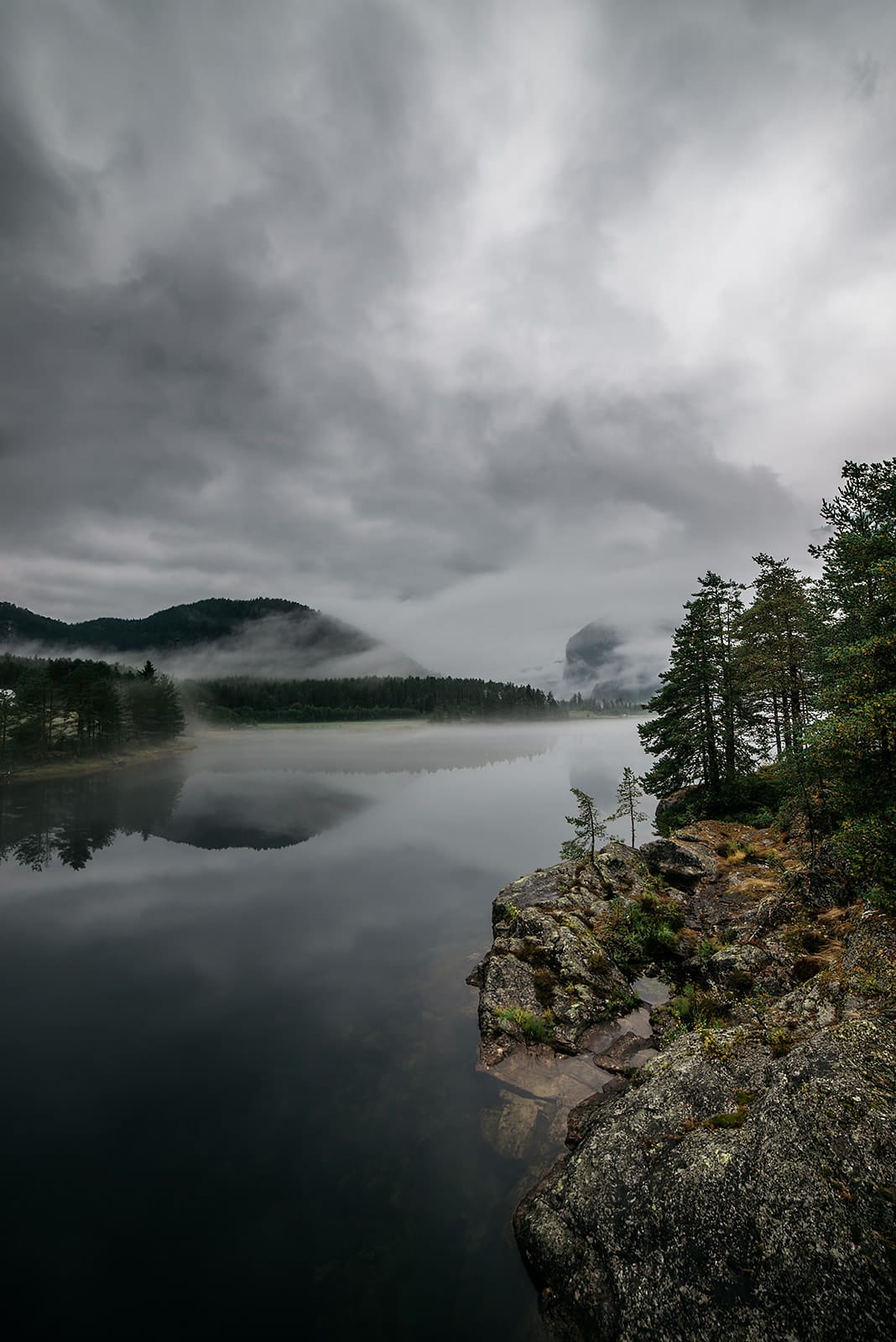 A misty lake bordered by rocky shores and pine trees reflects fog-covered hills and a cloudy sky in a tranquil Norwegian setting.