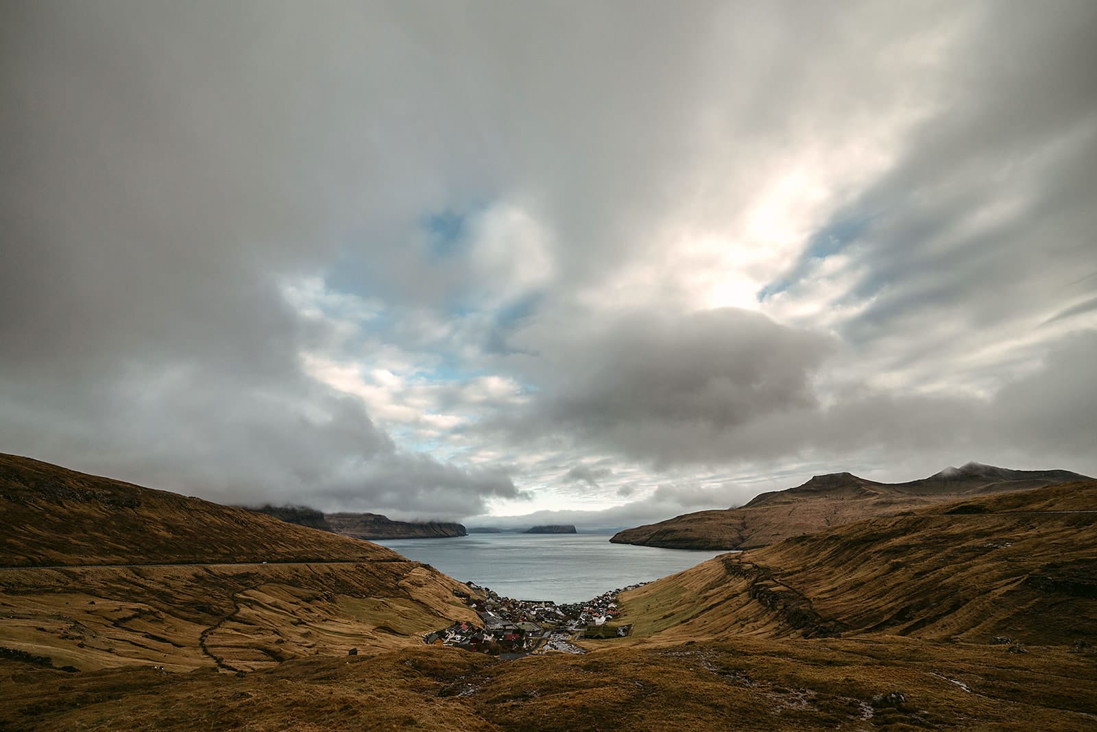 A small village nestled in a valley between rolling brown hills, leading down to a calm body of water under a dramatic sky with thick clouds and patches of blue, taken on the Faroe Islands