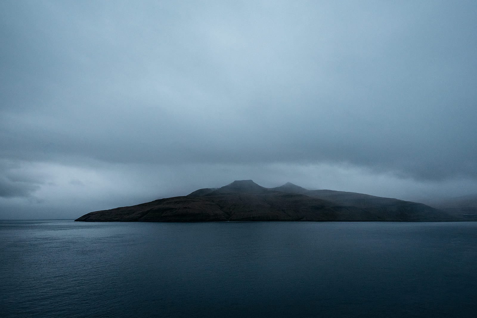 Dark, mist-covered island with gentle hills surrounded by calm, deep blue water under a heavy, overcast sky, taken on the Faroe Islands