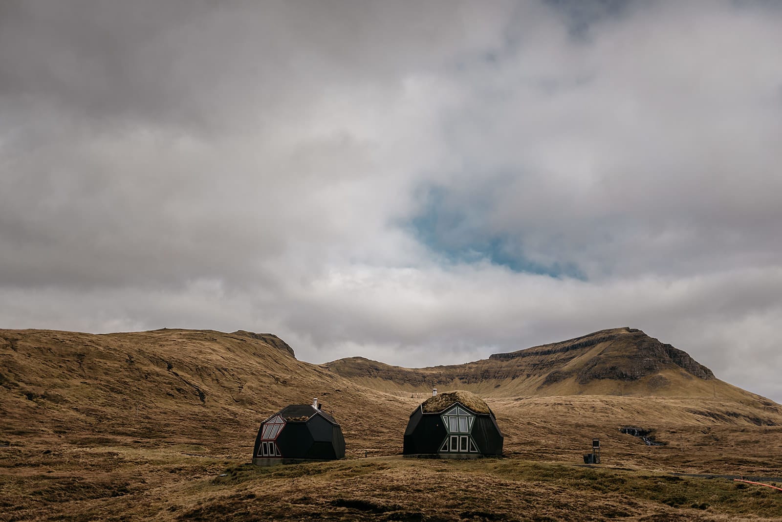 Two small, black, dome-shaped houses with grass-covered roofs sit on a grassy plain surrounded by rolling brown hills under a cloudy sky with patches of blue, taken on the Faroe Islands.