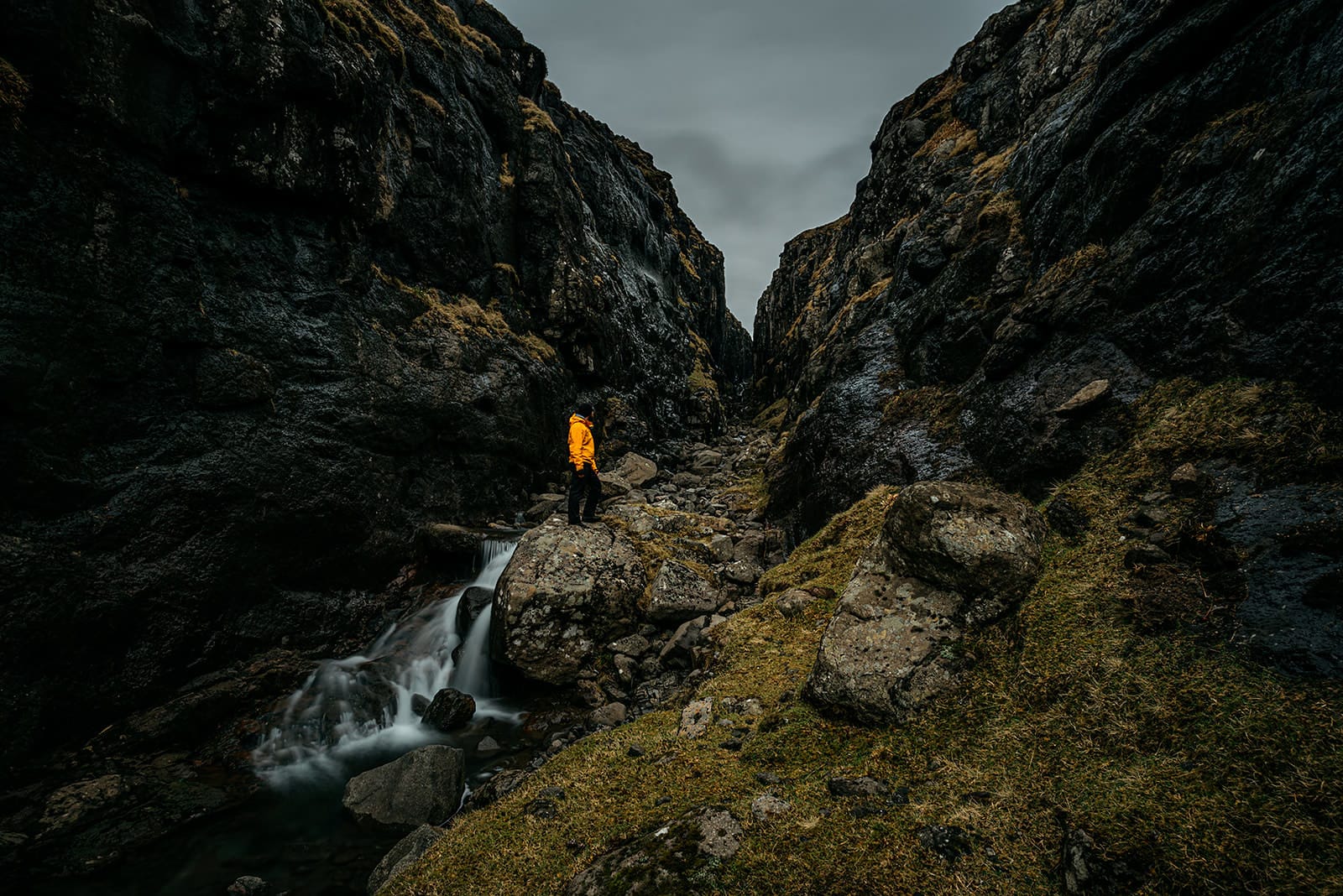 A person wearing a bright yellow jacket stands on a large rock beside a small cascading stream flowing through a narrow, rocky gorge with steep dark cliffs covered in patches of moss, taken on the Faroe Islands.