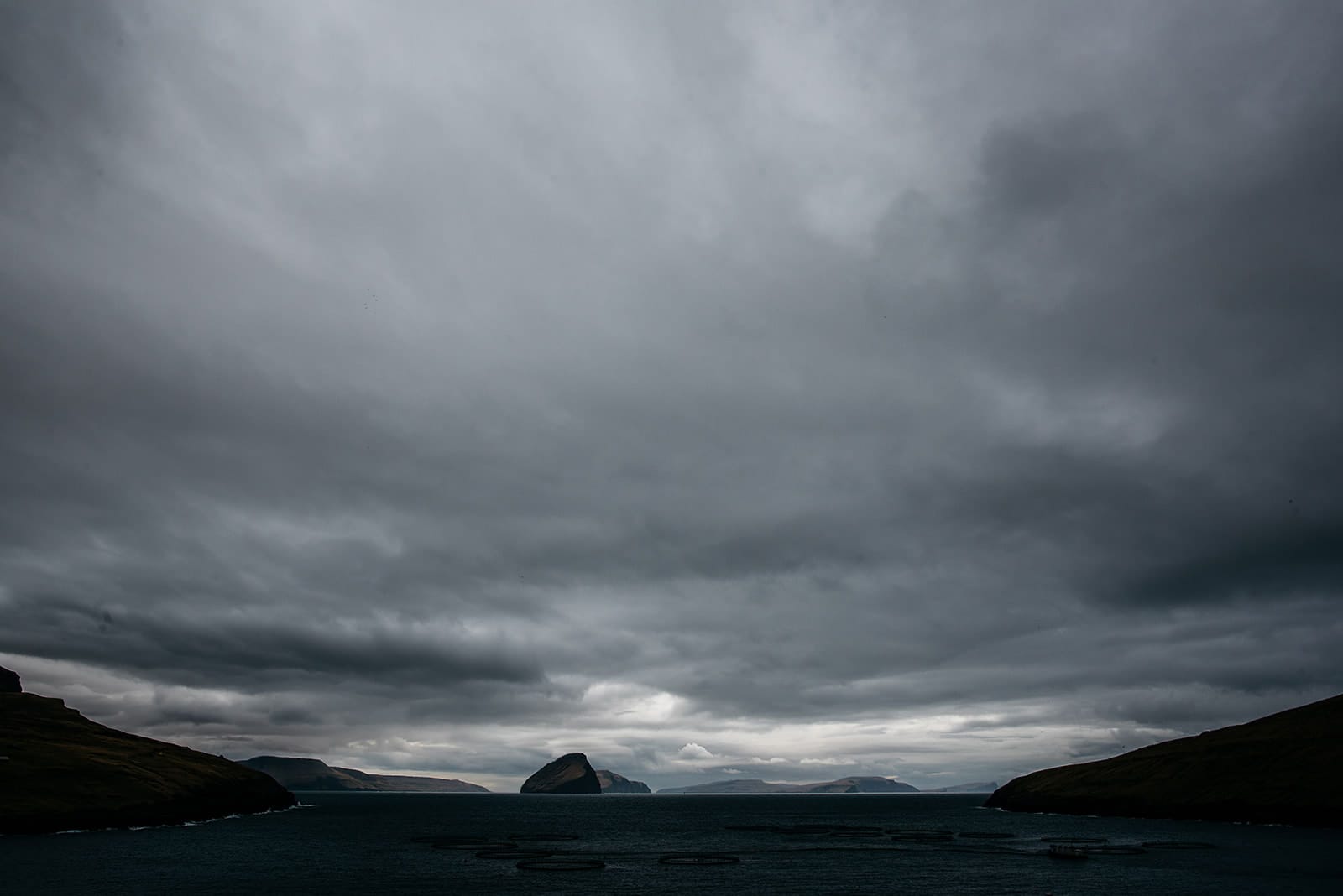 A dark, cloudy sky hangs over a calm sea with several distant islands, framed by shadowed hills on either side and circular fish farming pens visible in the water, taken on the Faroe Islands.
