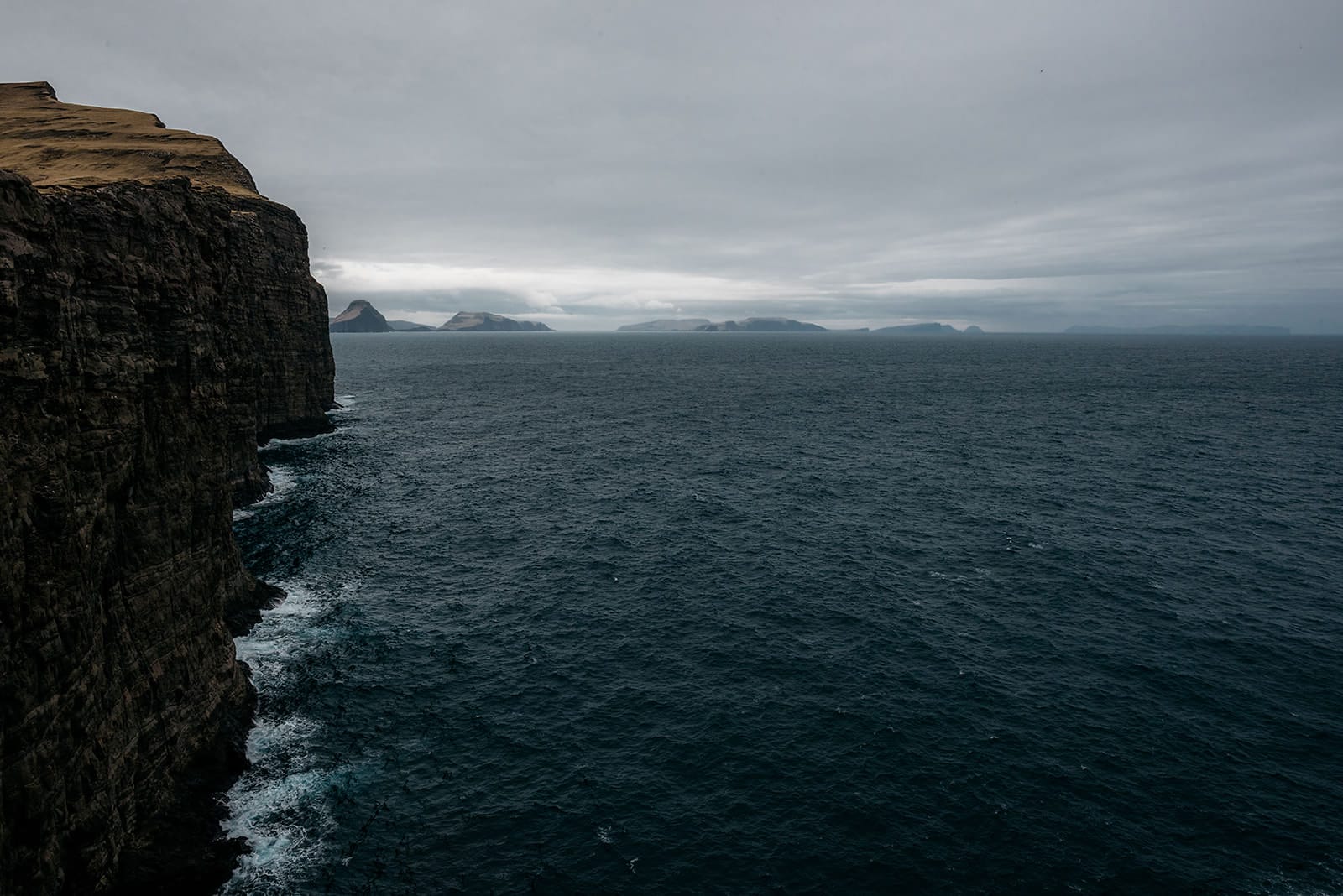 Steep, dark cliffs drop sharply into a deep blue, choppy sea with distant islands visible under a gray, overcast sky, taken on the Faroe Islands.
