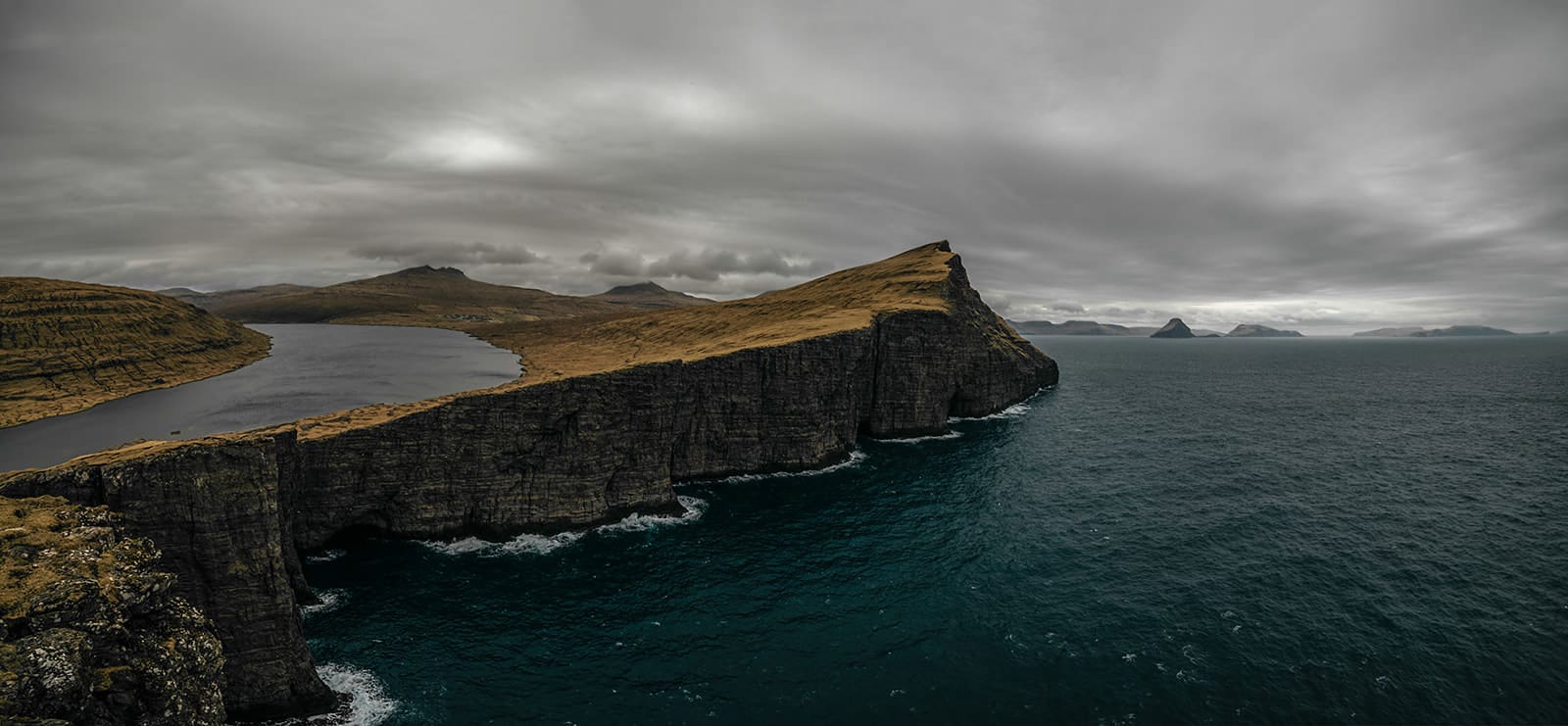 A dramatic cliff with steep, dark rock faces and a grassy top extends into a deep blue sea, with a calm lake and rolling brown hills behind it under a cloudy gray sky, taken on the Faroe Islands.
