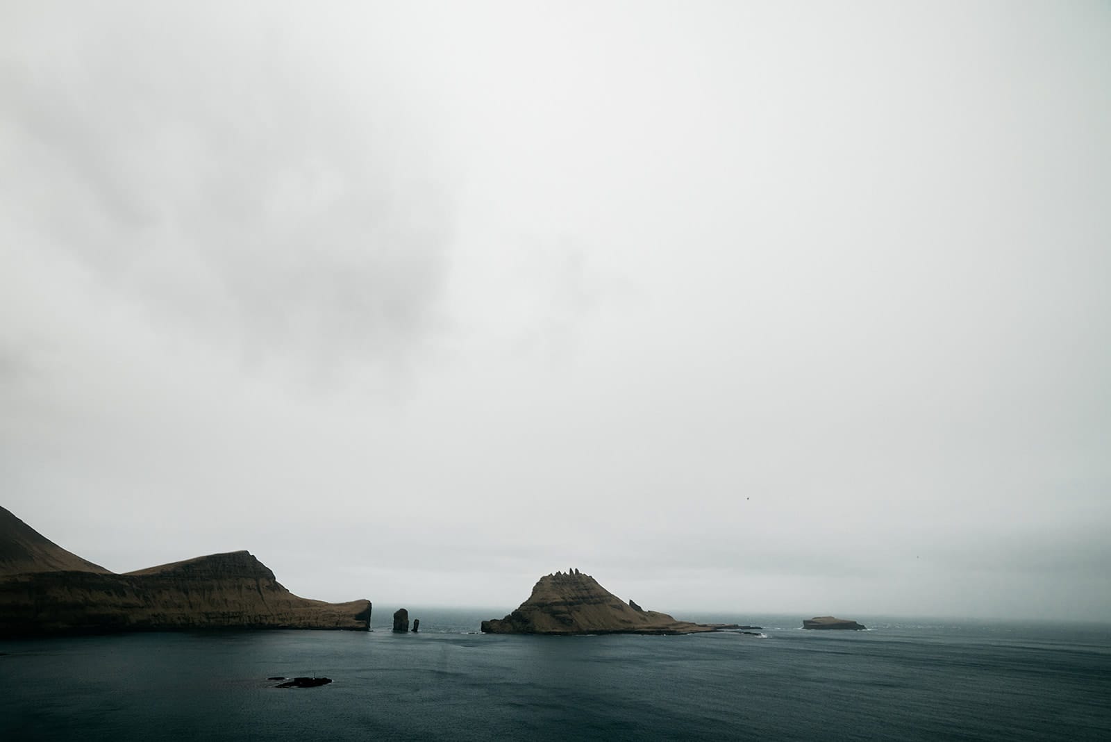A group of small, rocky islands with steep, brown cliffs rises from a calm, dark sea under a pale, overcast sky, taken on the Faroe Islands.