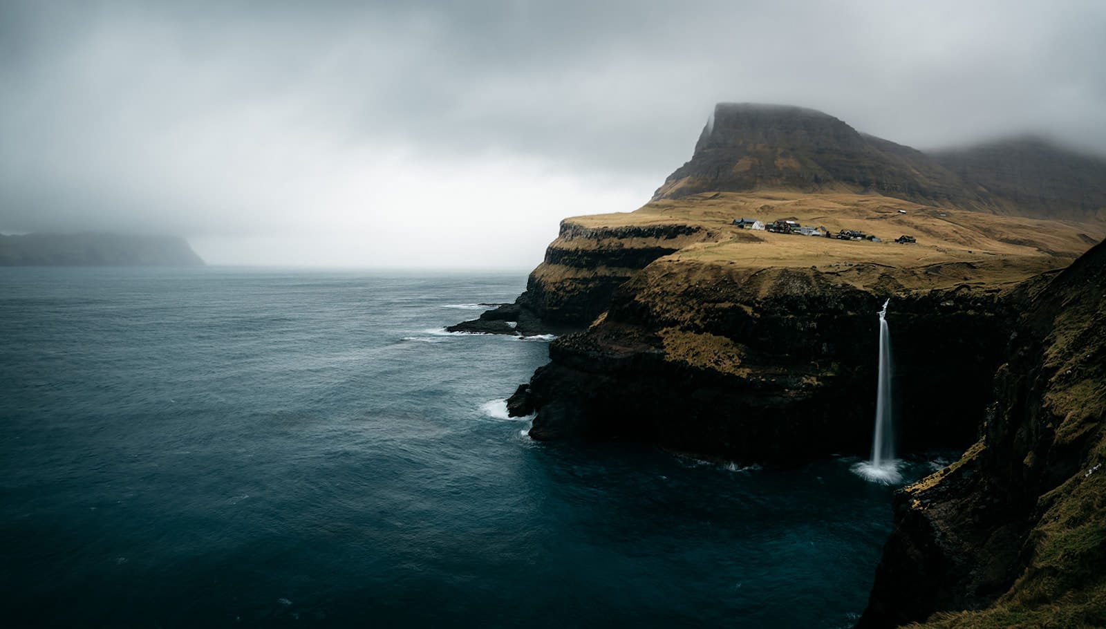 Remote coastal cliff with a tall waterfall plunging into a dark blue ocean, topped by a small village under a cloudy, misty sky, taken on the Faroe Islands