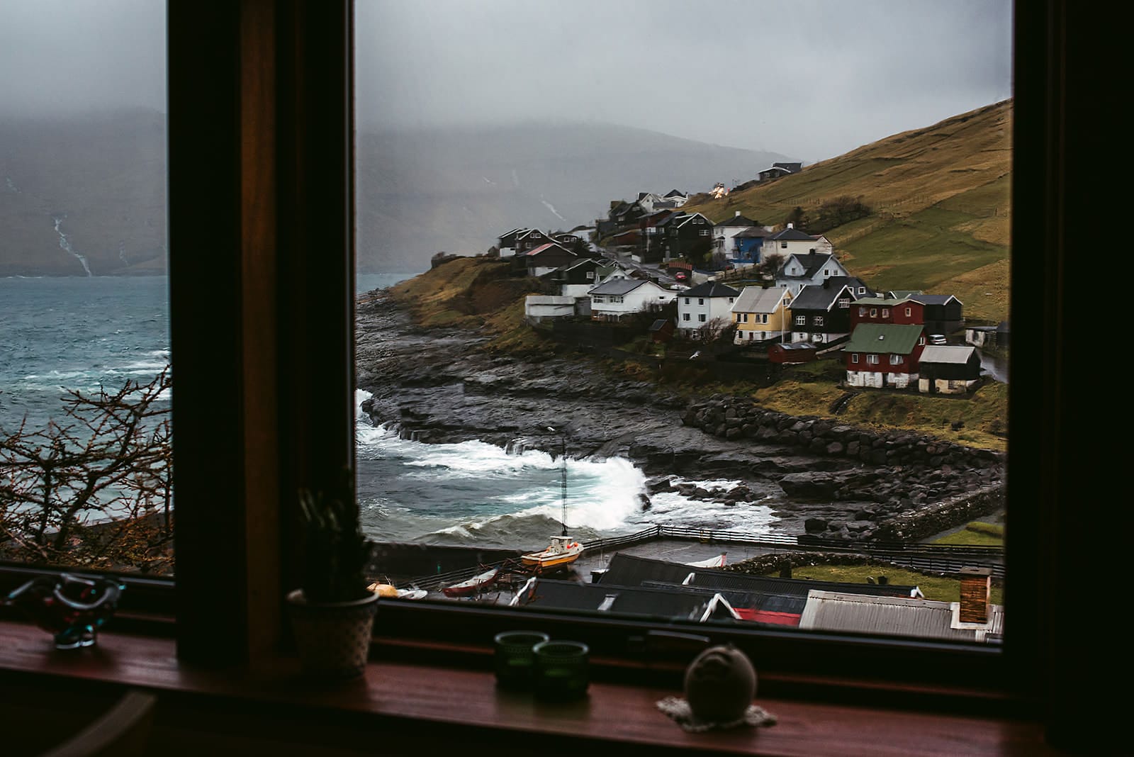 View through a window overlooking a small coastal village with colorful houses perched on rocky shores beside a turbulent sea, with misty hills in the background, taken on the Faroe Islands.