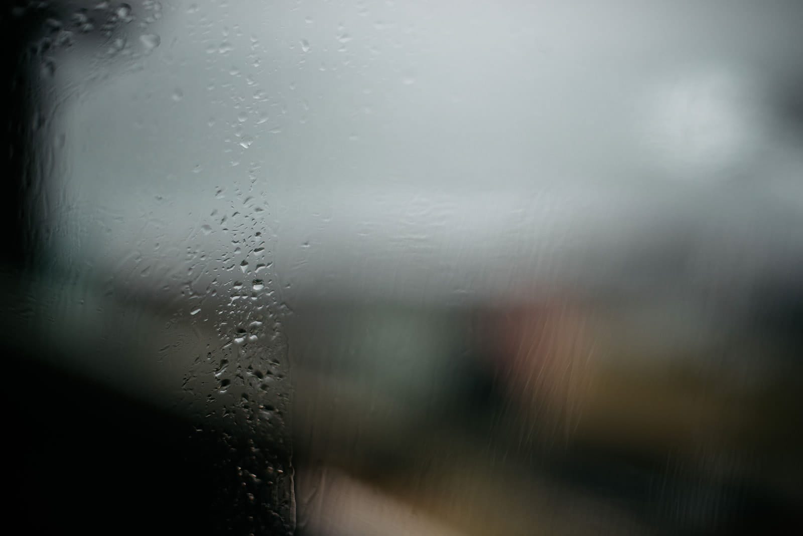 Raindrops streak down a windowpane, blurring the view of an indistinct, gray, and overcast landscape beyond.