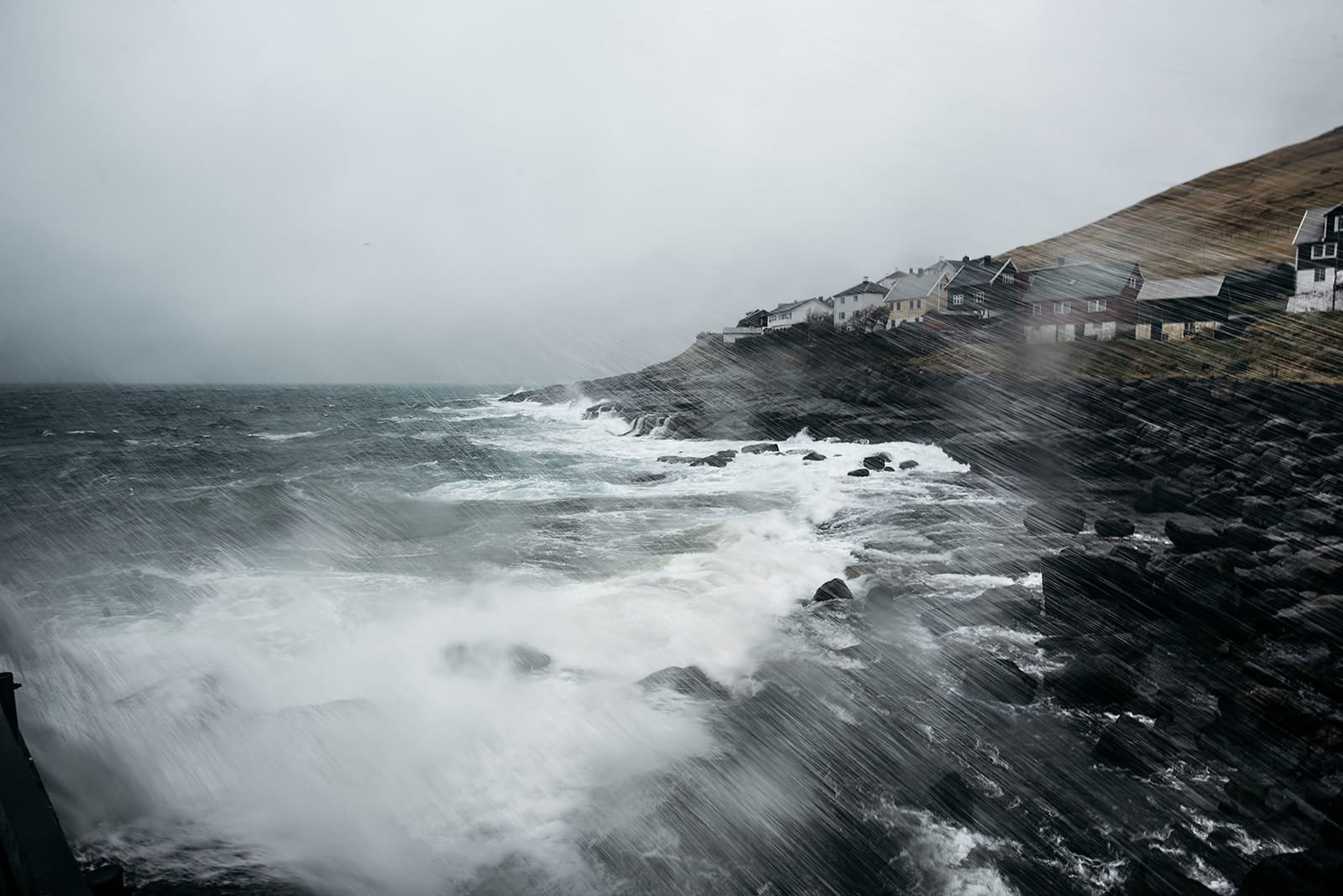 Strong rain and waves crash against a rocky shoreline with houses perched on a steep hill under a gray, stormy sky, taken on the Faroe Islands.