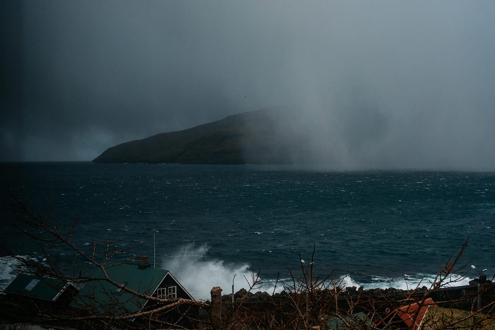 Waves crash against a coastal house with a green roof under a dark, stormy sky, while a misty island looms in the background, taken on the Faroe Islands.