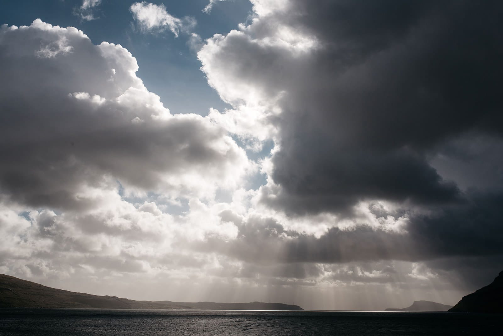 Sunlight breaks through dramatic dark clouds, casting rays over a calm sea framed by distant hills, taken on the Faroe Islands.