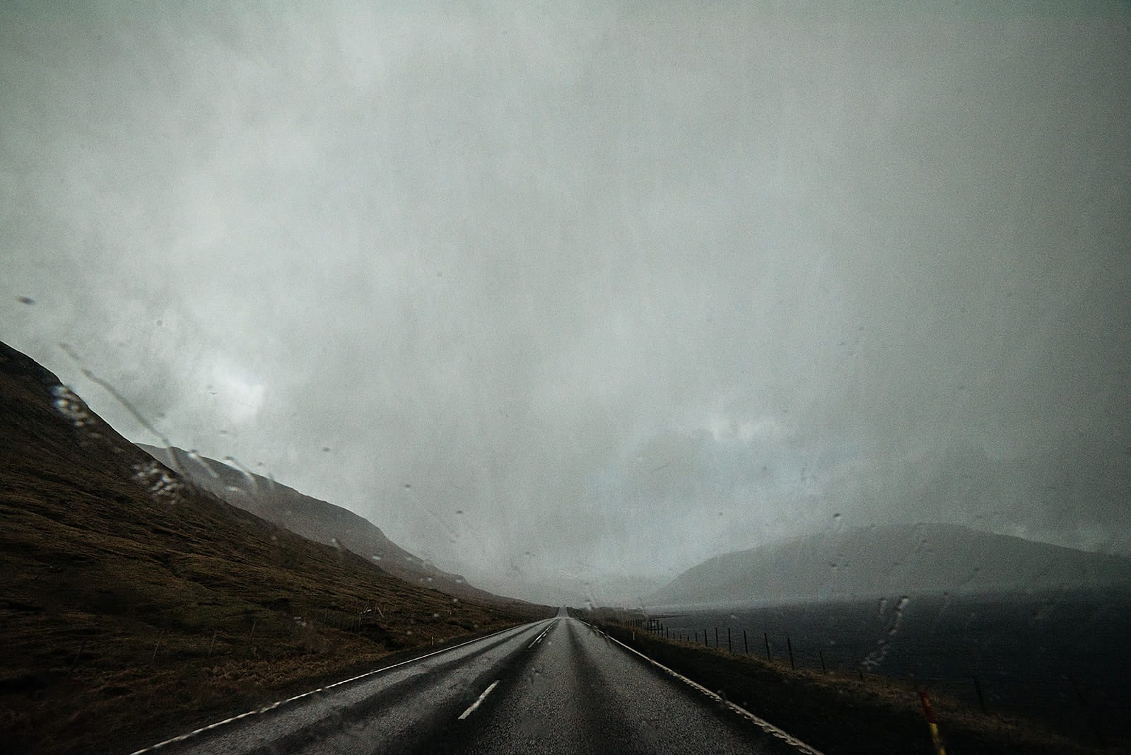A wet road stretches straight ahead between dark hills and a body of water under a heavy, gray sky with raindrops on the camera lens, taken on the Faroe Islands.