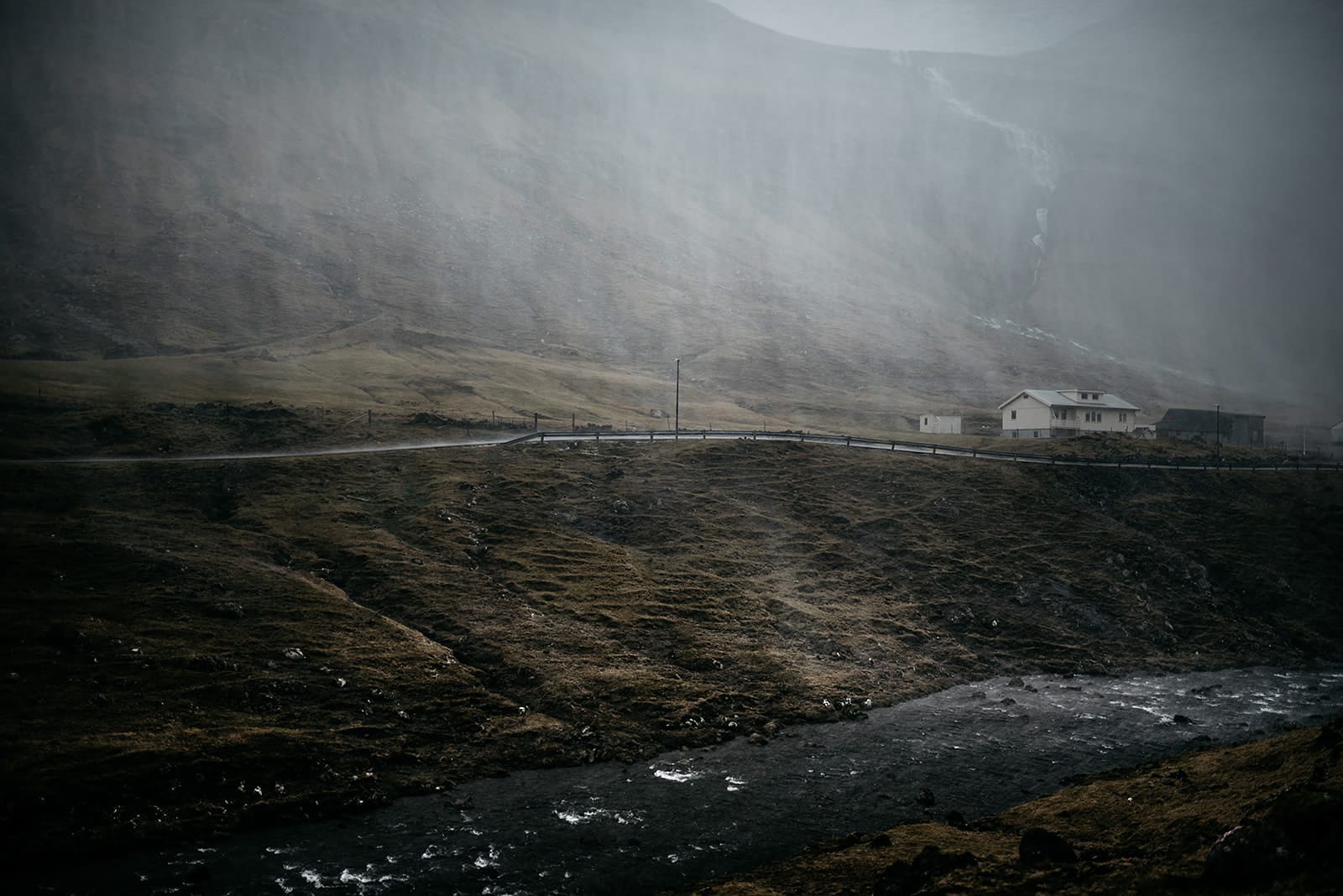 A misty scene with a small white house near a winding road beside a flowing stream, set against a steep, fog-covered hillside, taken on the Faroe Islands.