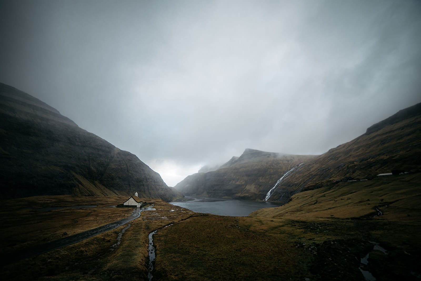 A solitary white church stands in a misty valley between steep hills with a waterfall cascading into a lake, taken on the Faroe Islands.