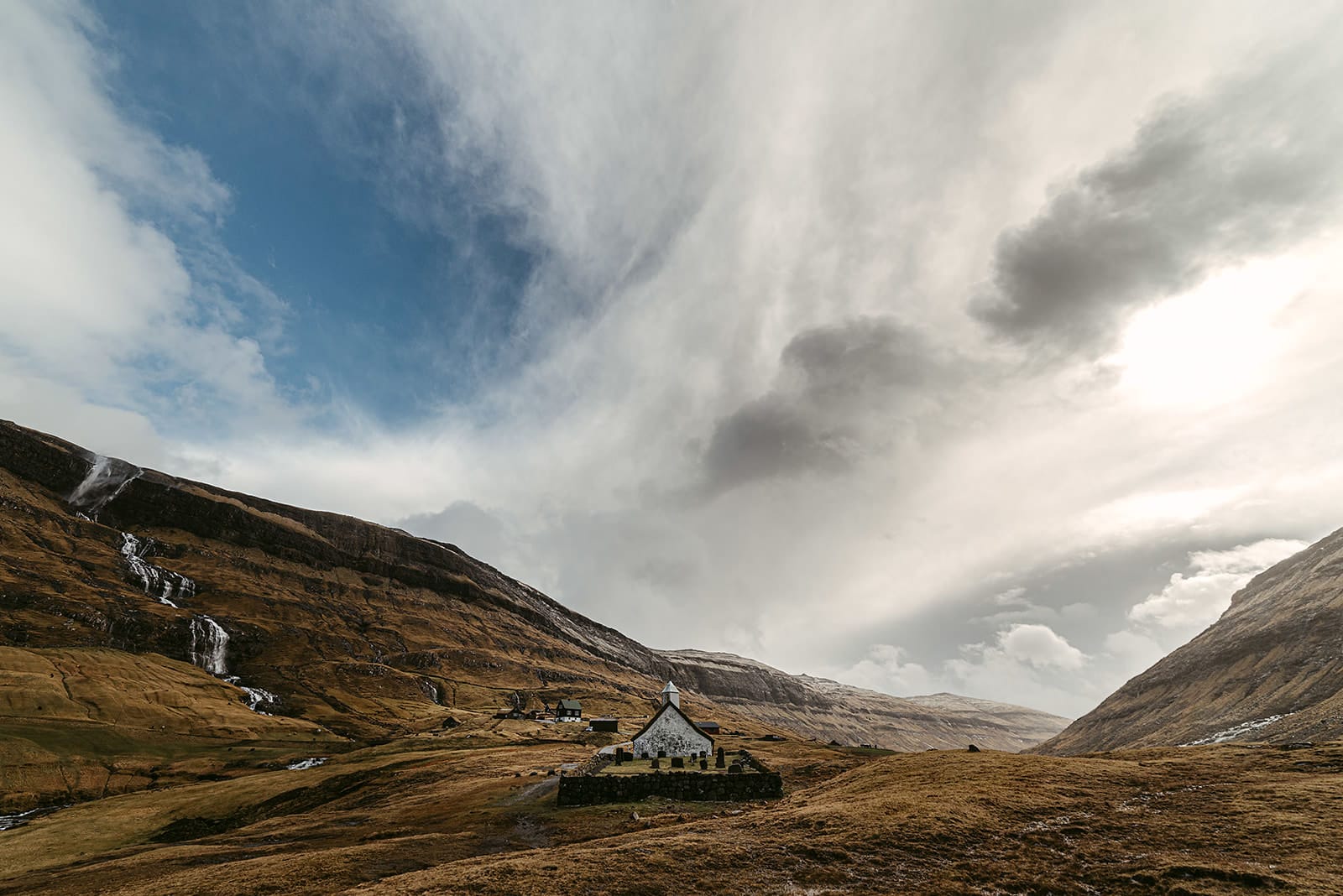 A white church sits in a sunlit valley surrounded by steep hills with waterfalls under a partly cloudy blue sky, taken on the Faroe Islands.