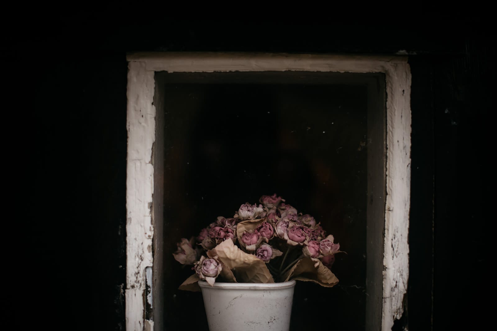 A small white pot holds a bouquet of dried pink roses behind a weathered window frame with peeling white paint, taken on the Faroe Islands.
