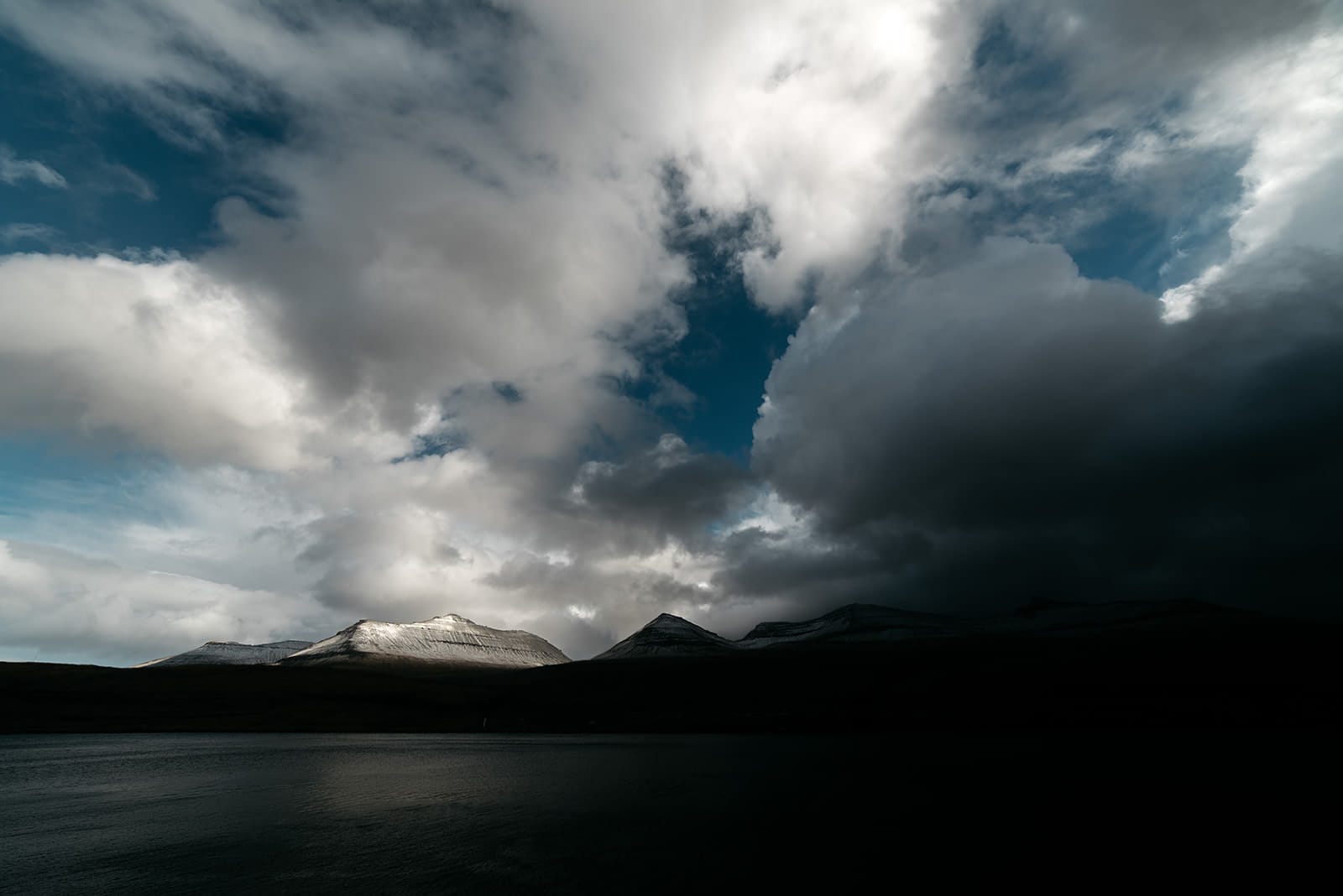 Sunlight breaks through dark clouds to illuminate snow-capped mountain peaks above a shadowed lake, taken on the Faroe Islands.