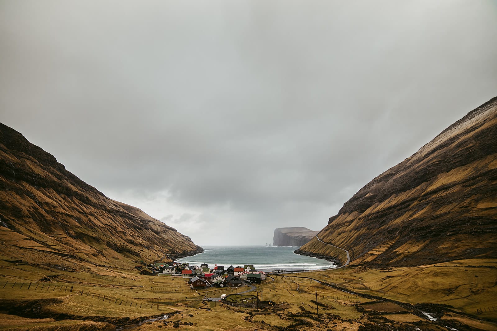 A small village with colorful houses nestles in a valley between steep hills leading to a narrow sea inlet under a cloudy sky, taken on the Faroe Islands.