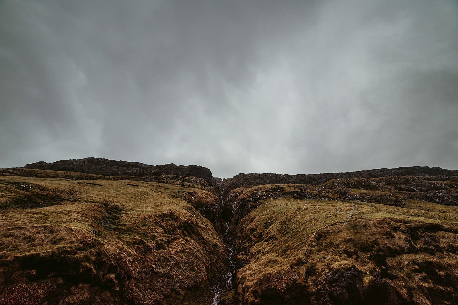A narrow stream cuts through steep, grassy hills under a heavy, overcast sky, taken on the Faroe Islands.