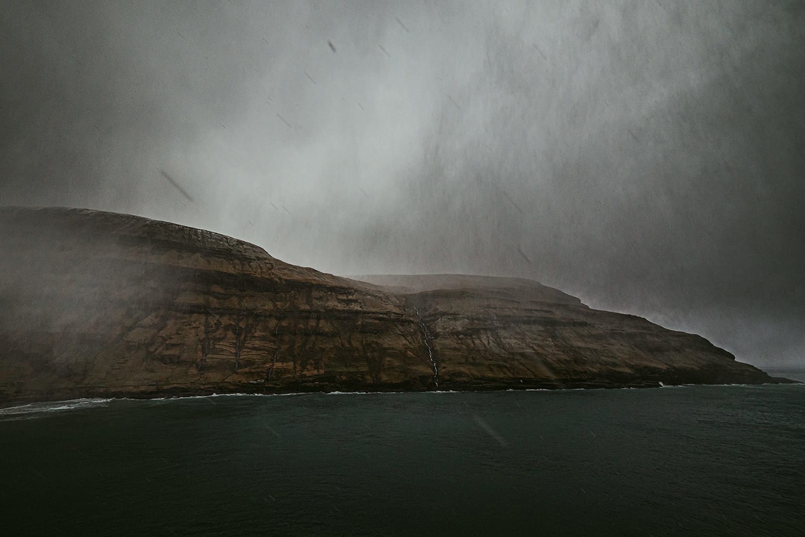 Heavy rain falls over steep, brown cliffs beside a dark sea under a stormy gray sky, taken on the Faroe Islands.