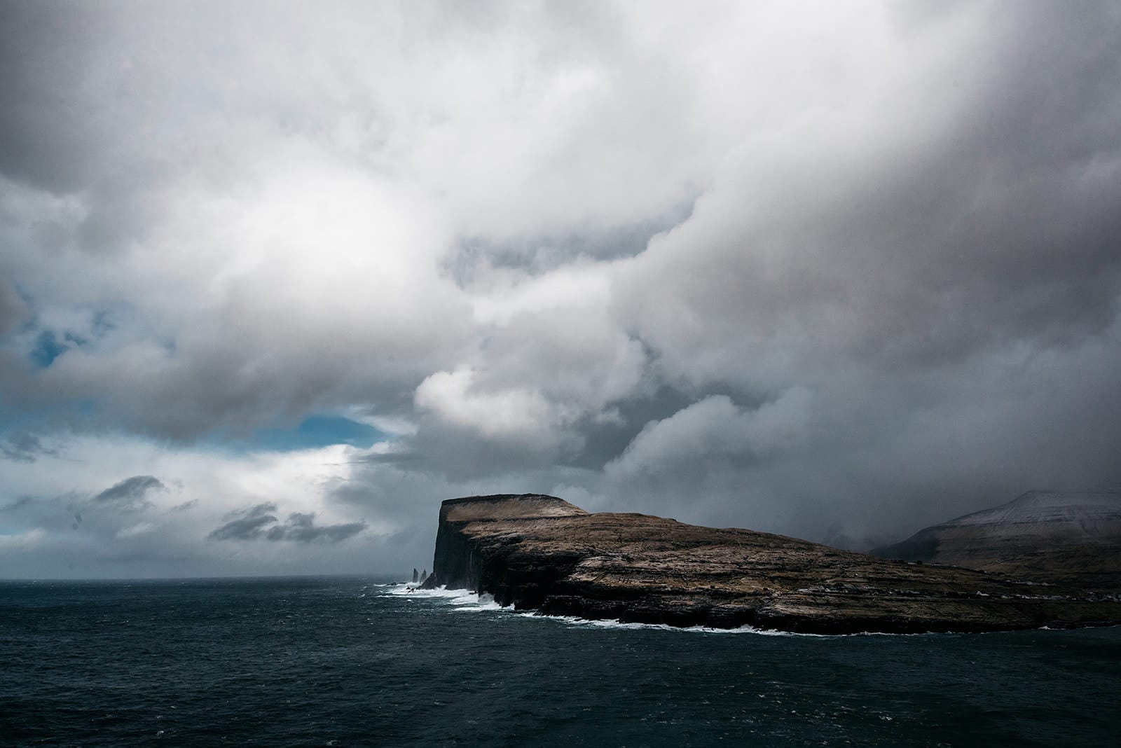 Waves crash against a rugged cliff jutting into a dark sea beneath a dramatic sky of heavy clouds and patches of blue, taken on the Faroe Islands.