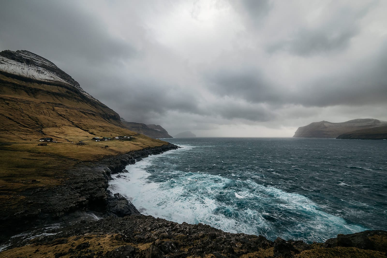 Waves crash against rocky cliffs along a rugged coastline with scattered houses under a cloudy, gray sky, taken on the Faroe Islands.