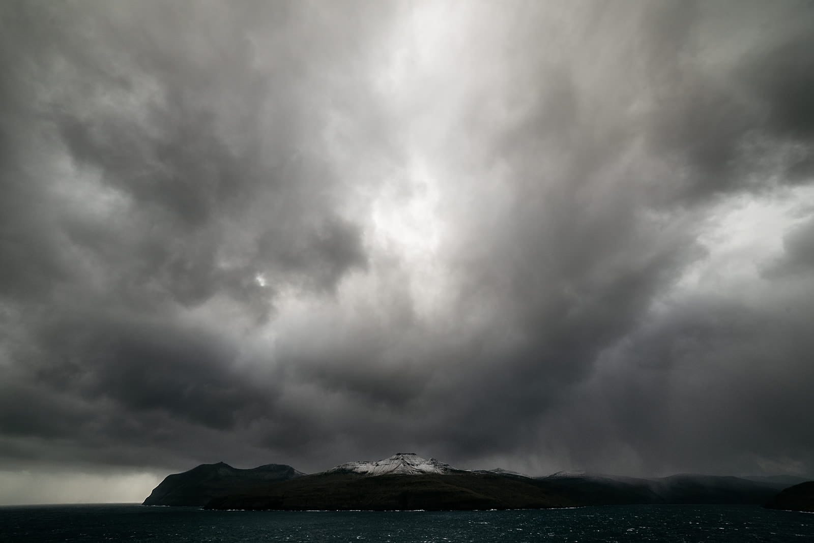 Dark storm clouds gather over a snow-capped mountain island surrounded by a turbulent sea, taken on the Faroe Islands.
