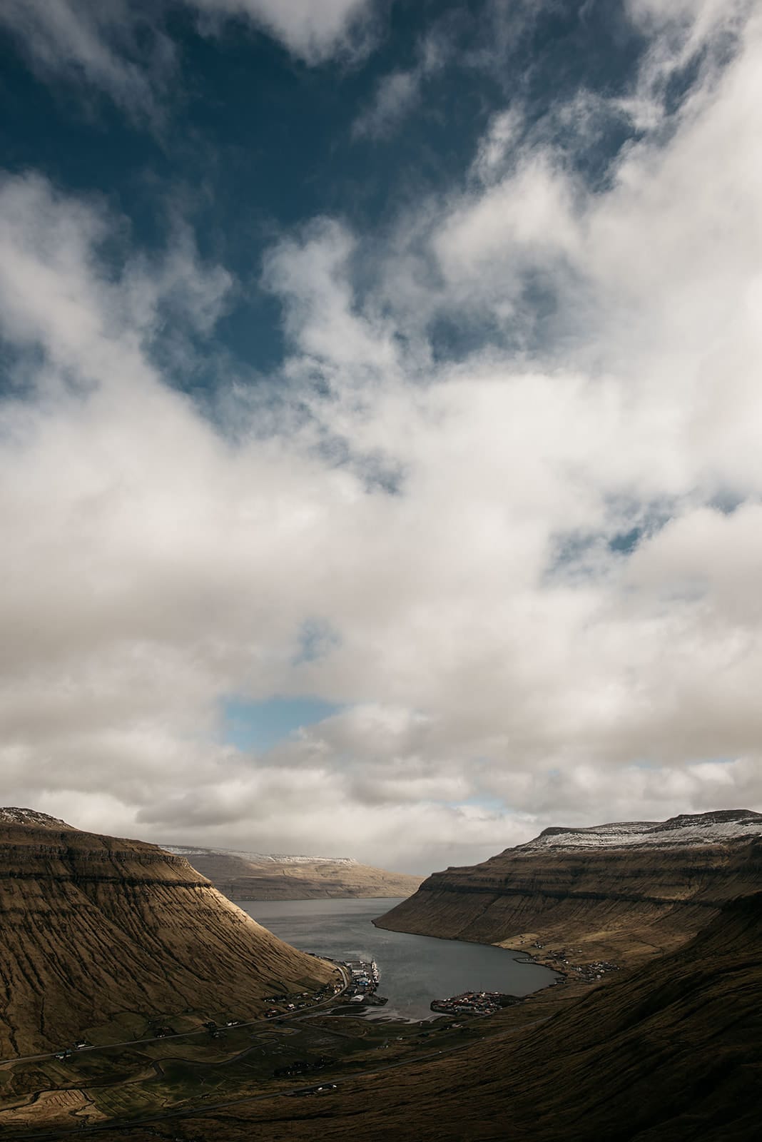 A fjord winds between steep, grassy mountains with small villages along the shore under a partly cloudy blue sky, taken on the Faroe Islands.