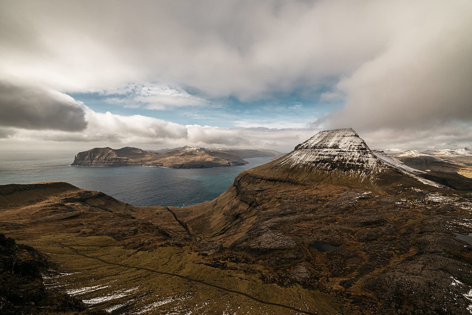 A snow-dusted, pyramid-shaped mountain overlooks a blue sea and distant islands under a partly cloudy sky, taken on the Faroe Islands.