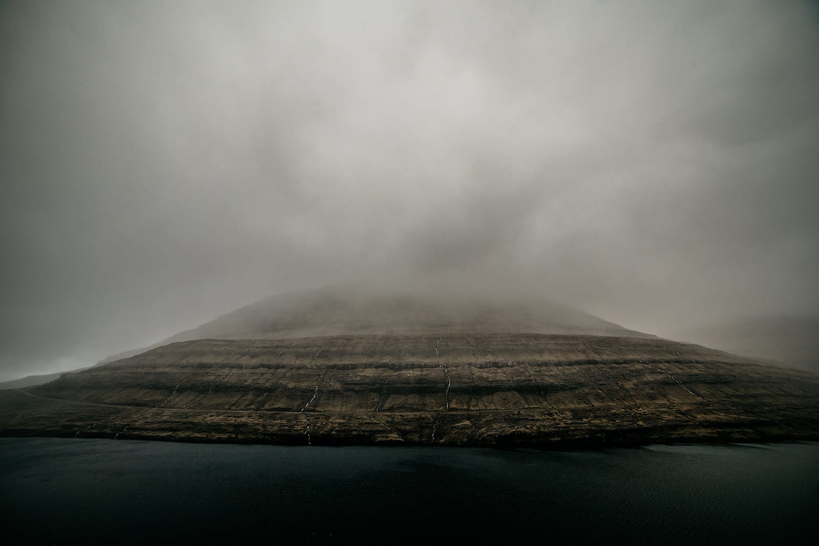 A large, layered mountain partially obscured by thick fog rises above a dark body of water, taken on the Faroe Islands.
